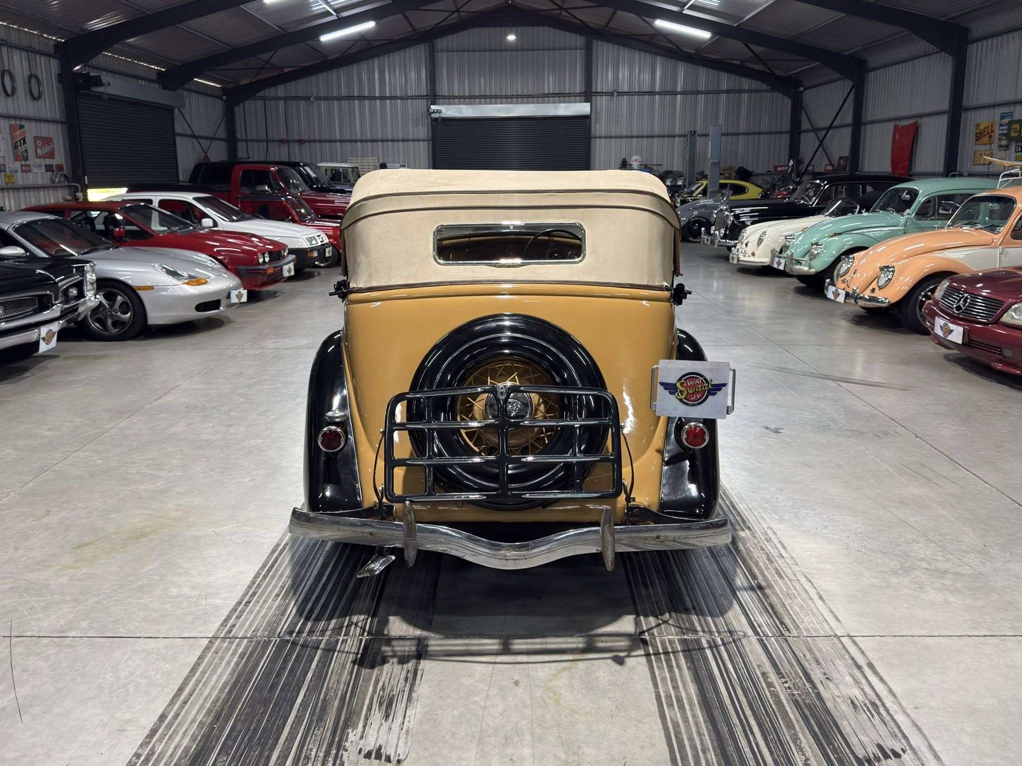 Rear view of a tan vintage car with a soft top in a classic car showroom, surrounded by other restored cars.