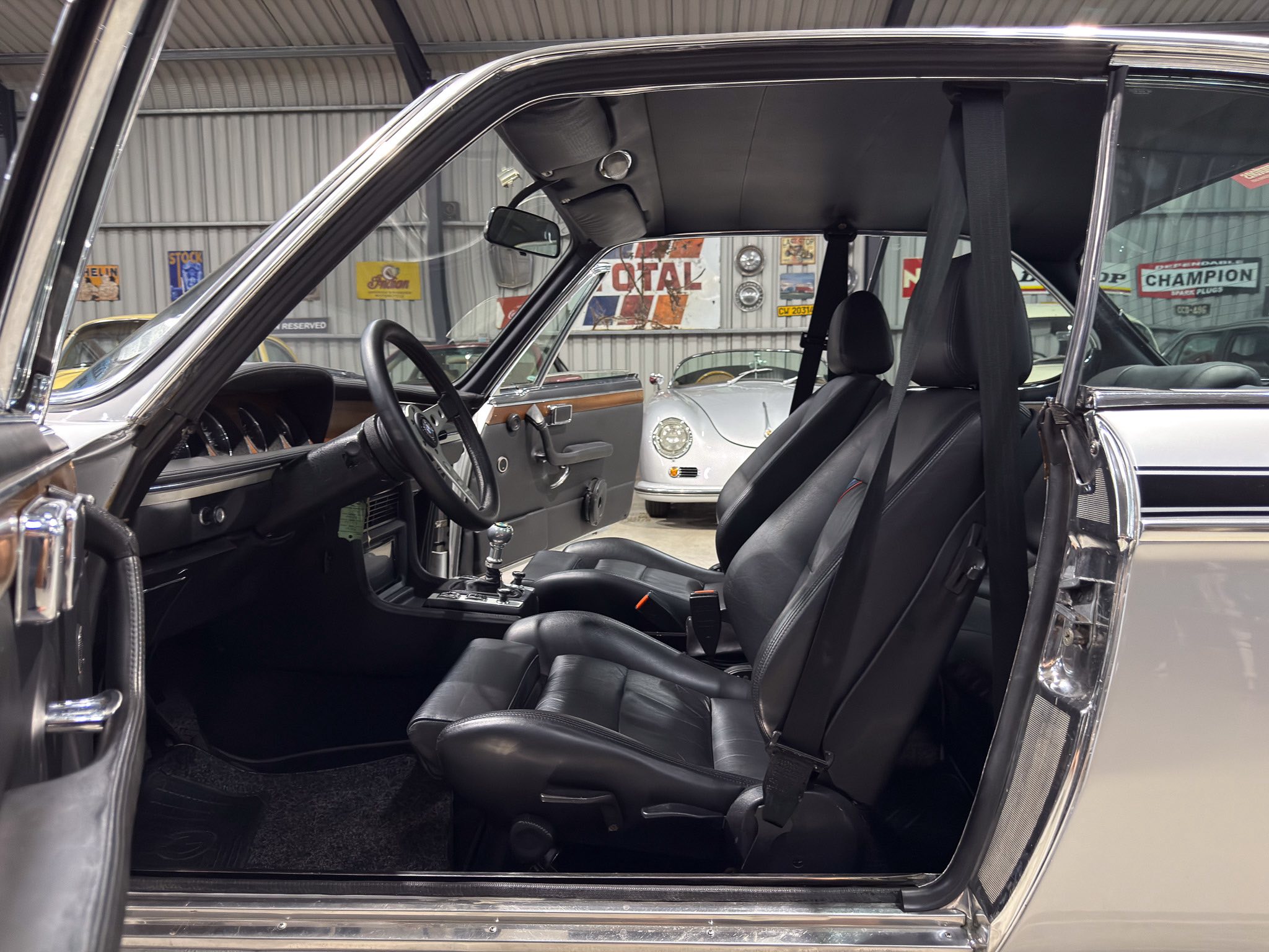 Black leather car interior with driver's seat and steering wheel visible in a classic showroom setting, other vintage cars seen through the windows behind.