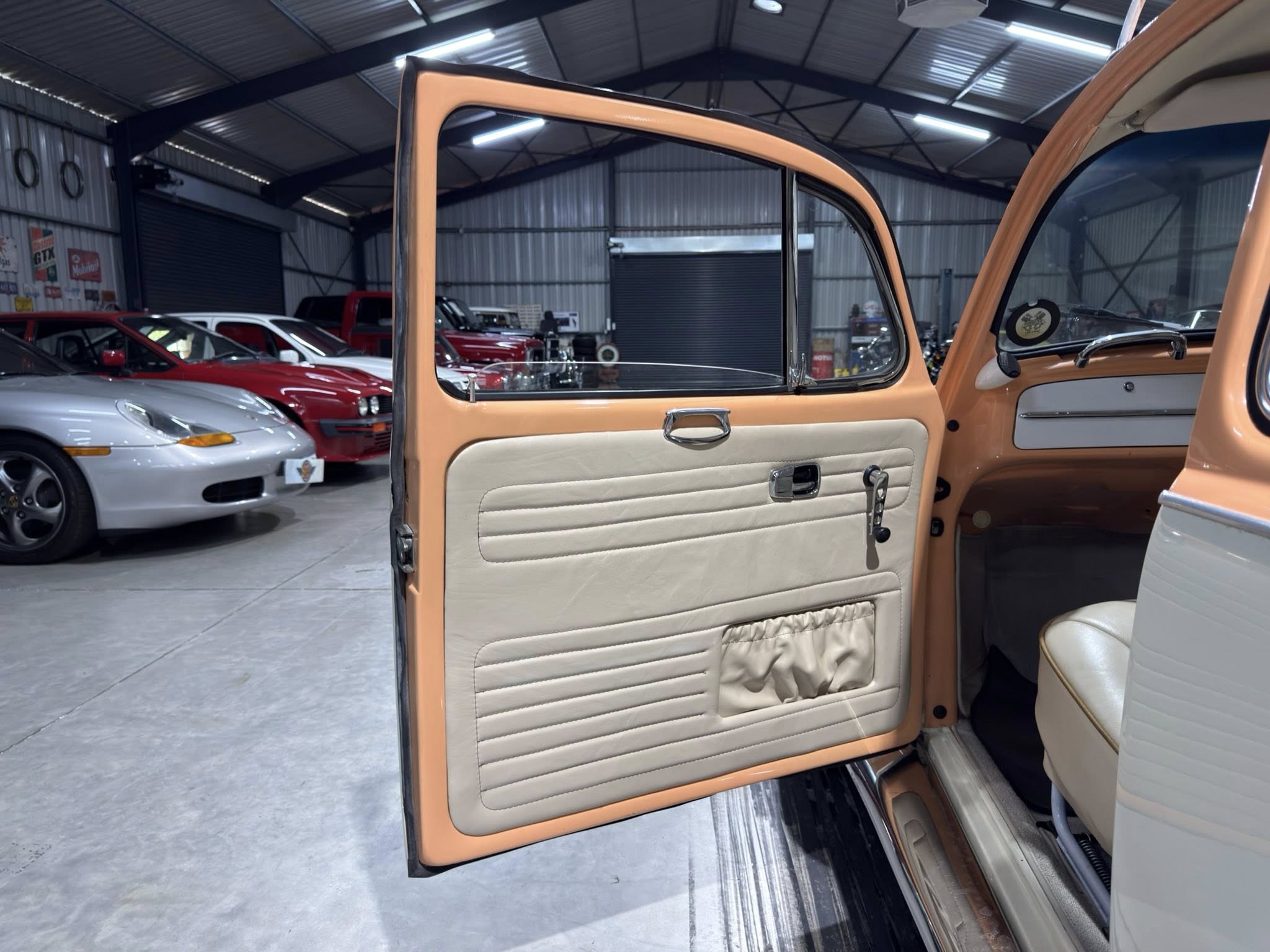 Open vintage car door showing tan interior panels inside a garage filled with classic cars and automotive tools.