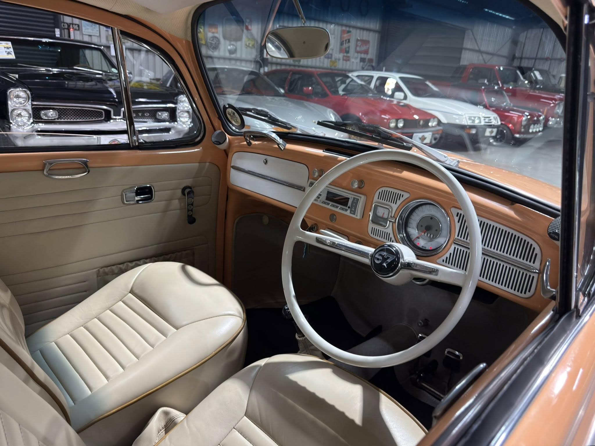 Interior of a vintage car with tan upholstery, white steering wheel, and chrome dashboard; a showroom full of classic cars visible outside the windows.