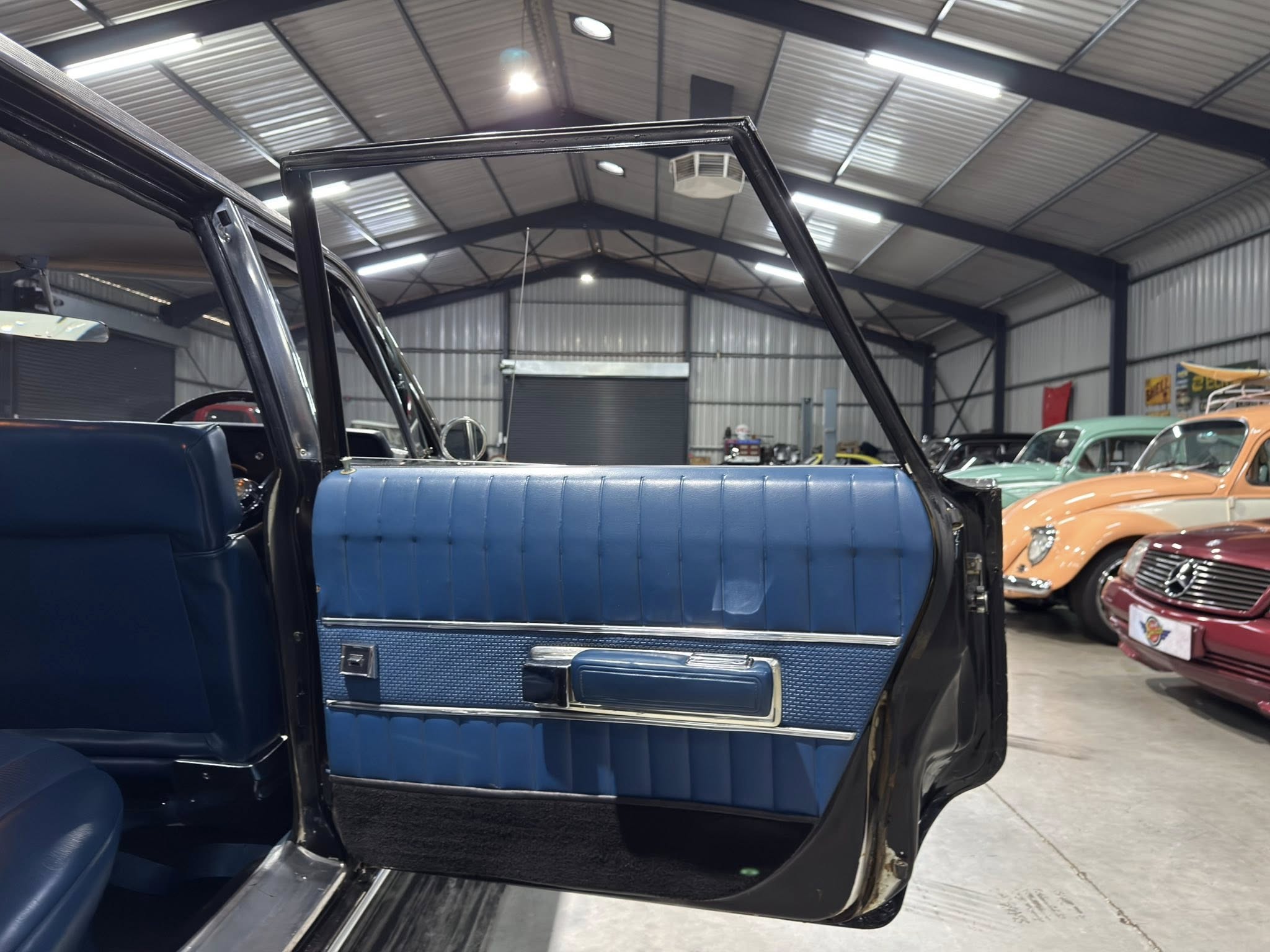 Open car door revealing blue interior upholstery in a showroom filled with vintage cars and a metal-arched ceiling in the background.
