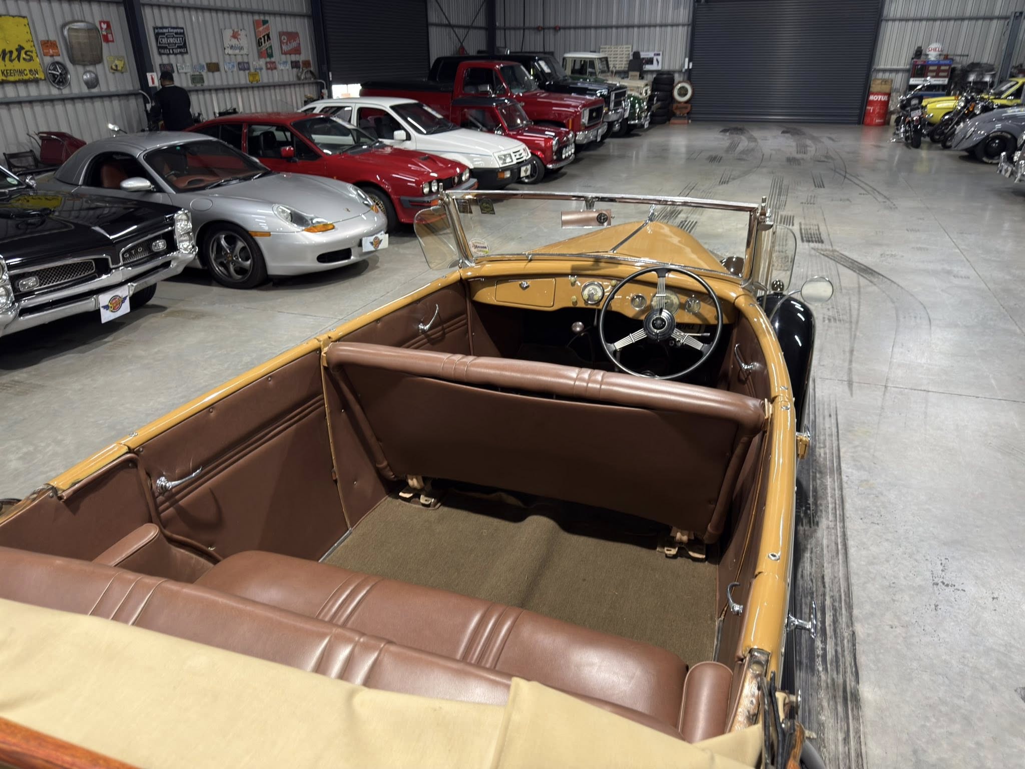 Inside a vintage car showroom with a row of classic cars lined up along a concrete floor, viewed from the tan convertible in the foreground.