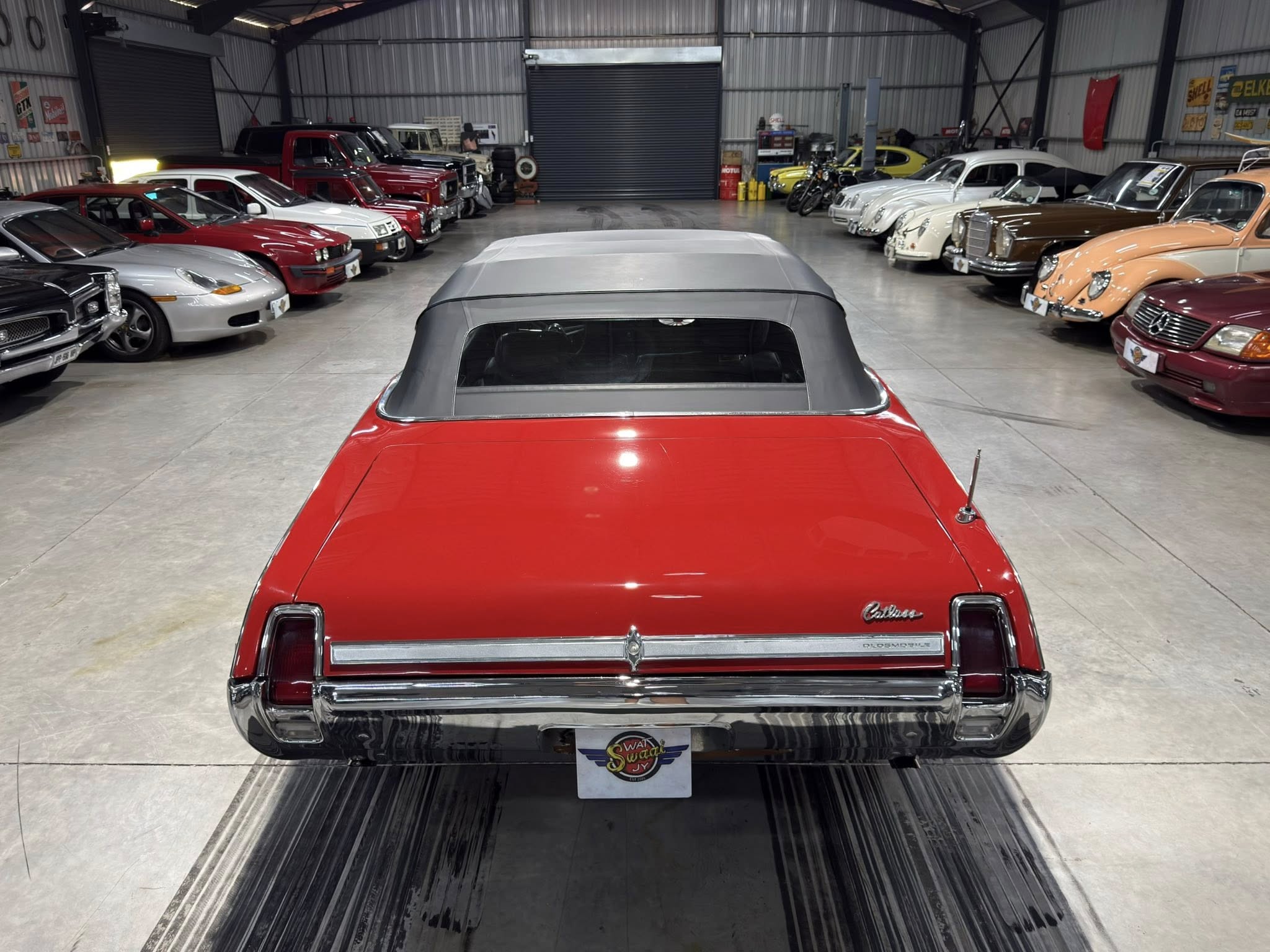 Rear view of a red convertible with a gray soft top, parked in a large indoor classic car showroom flanked by vintage cars on both sides.