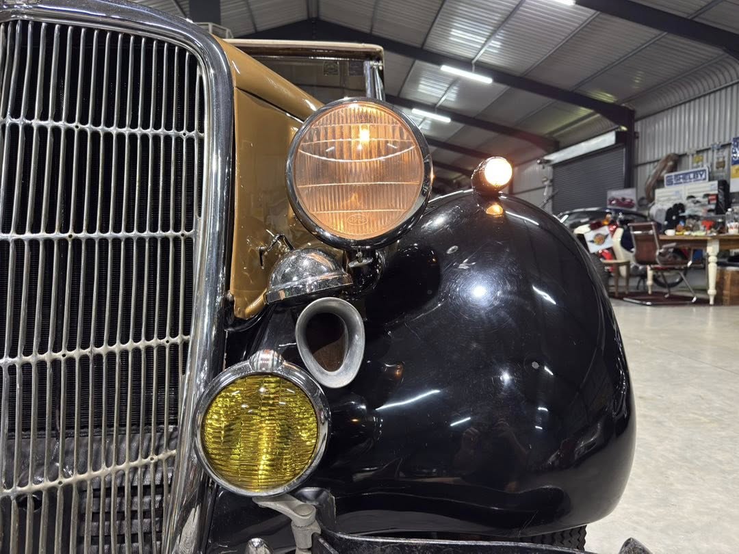 Close-up of a vintage black car front with a large round headlamp, yellow fog light, chrome grille, and tan body in a workshop showroom.