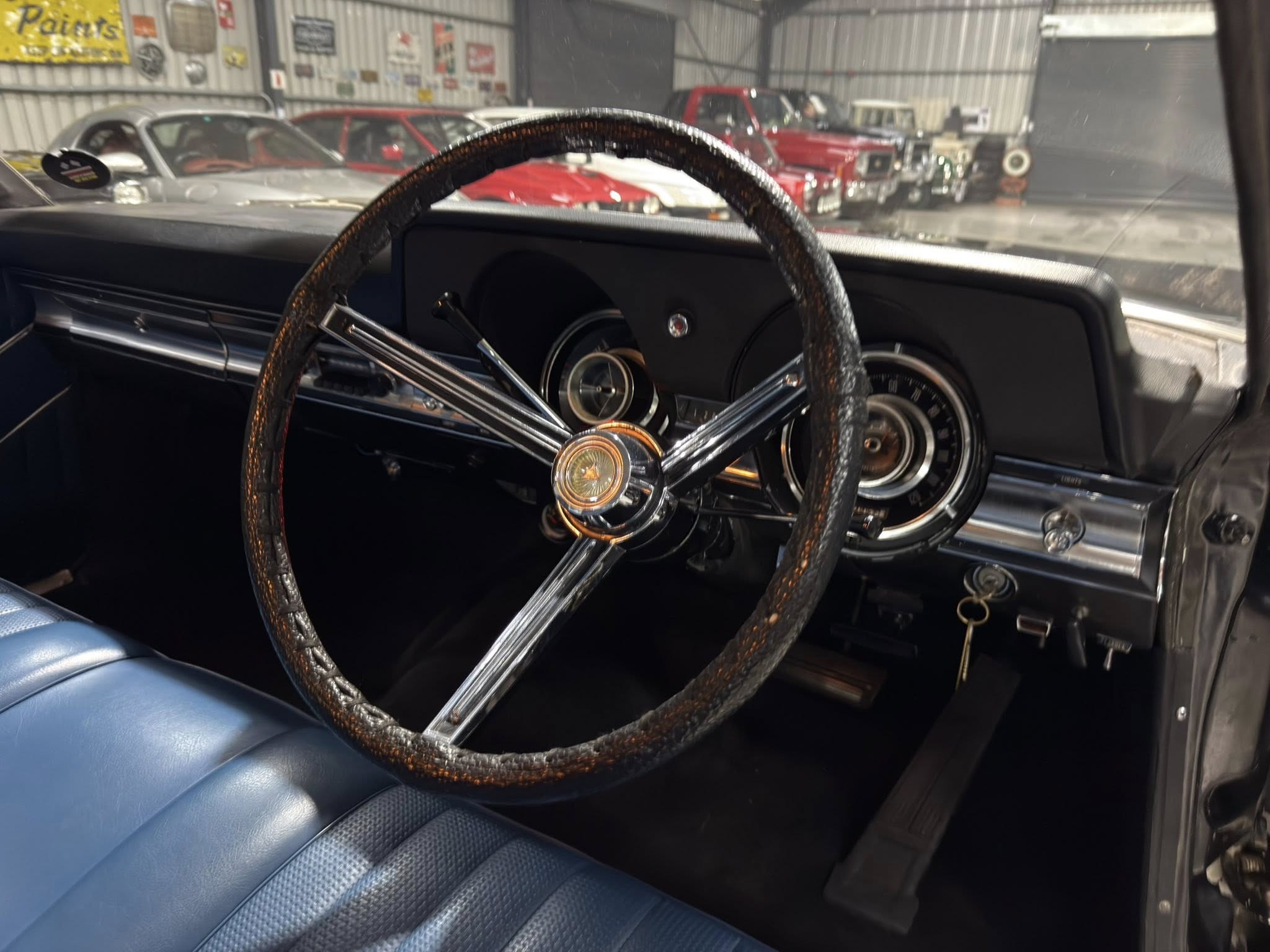 Interior of a vintage car showing a brown textured steering wheel and blue vinyl seats, with classic cars visible in a garage outside the windshield.