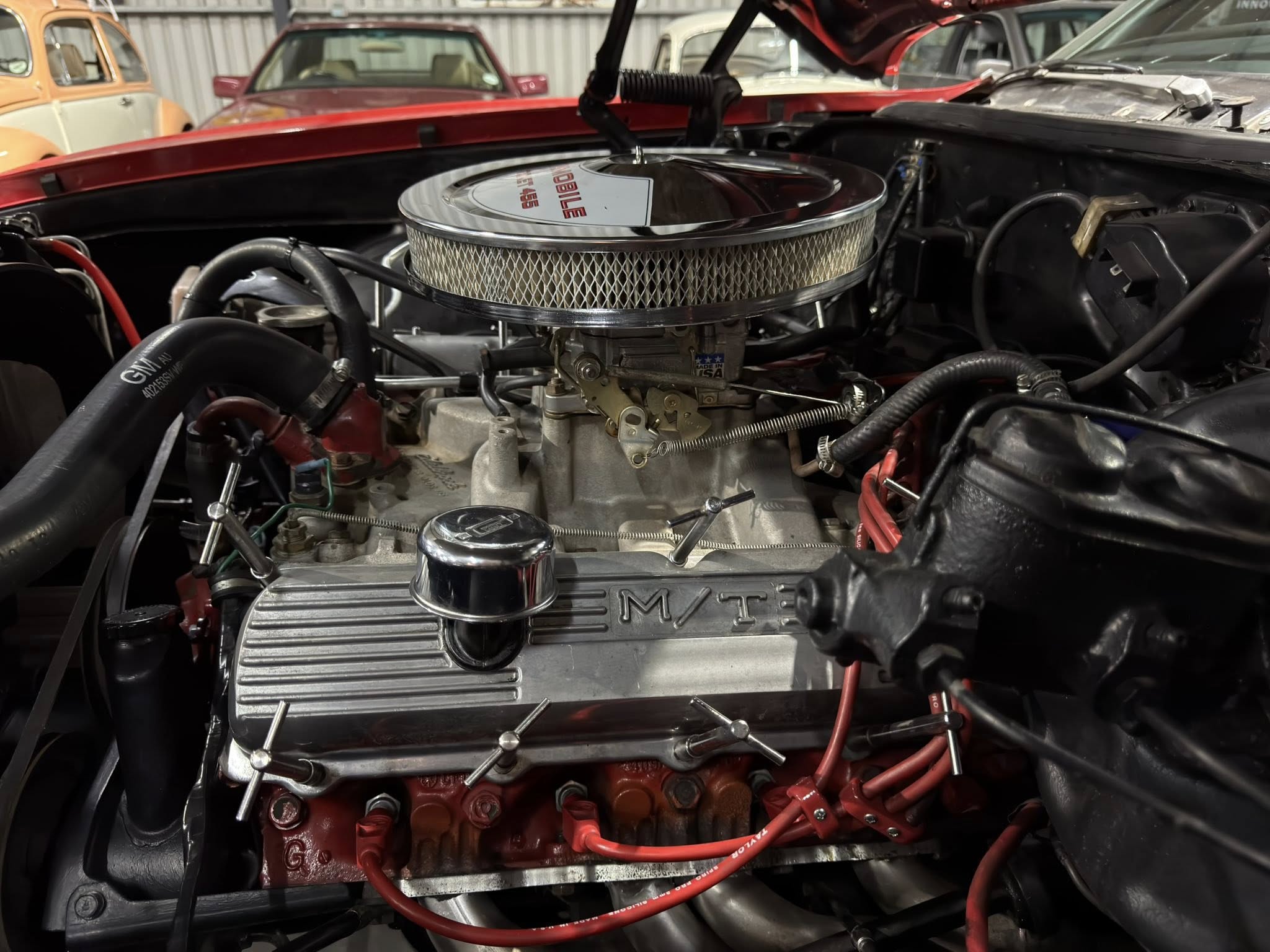 V8 car engine with chrome circular air filter housing and red ignition wires in a classic car restoration shop