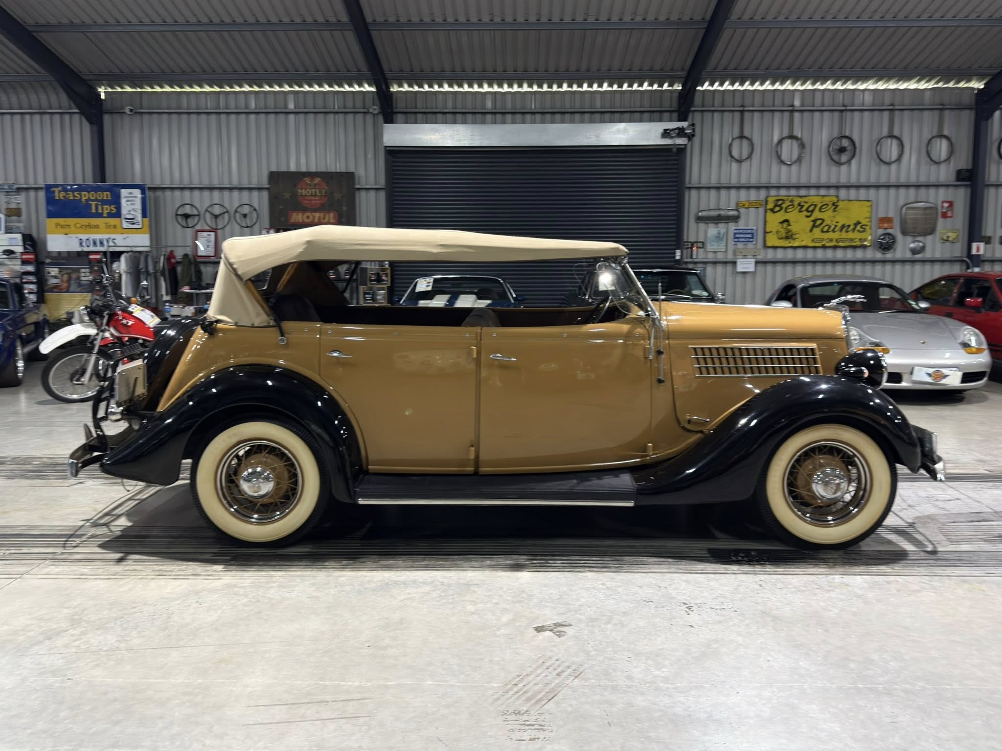 Beige vintage convertible with its soft top up inside a showroom-like garage, surrounded by classic cars and retro signs.