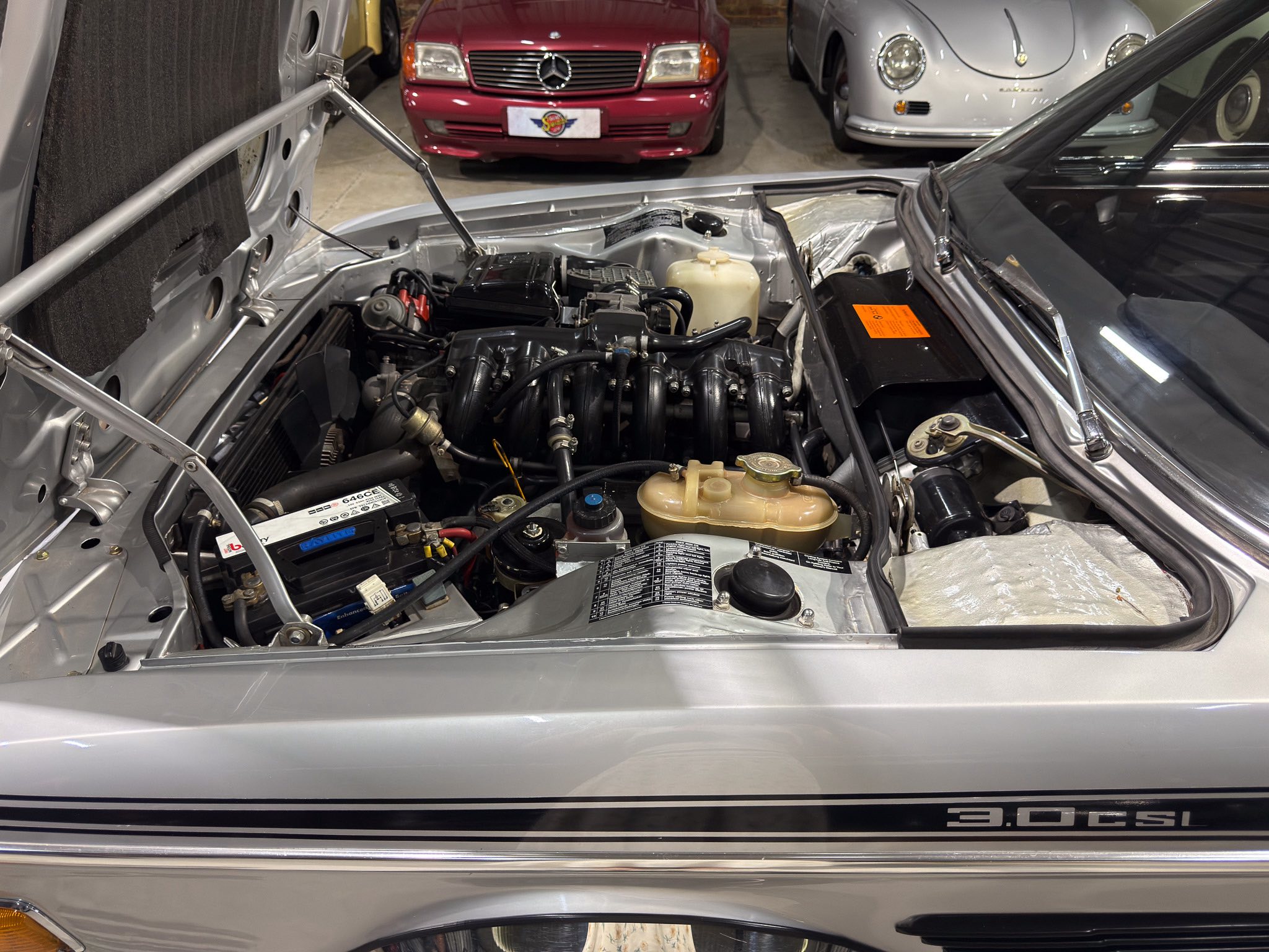 Open hood of a silver car showing its engine; showroom with red and silver classics in the background.",