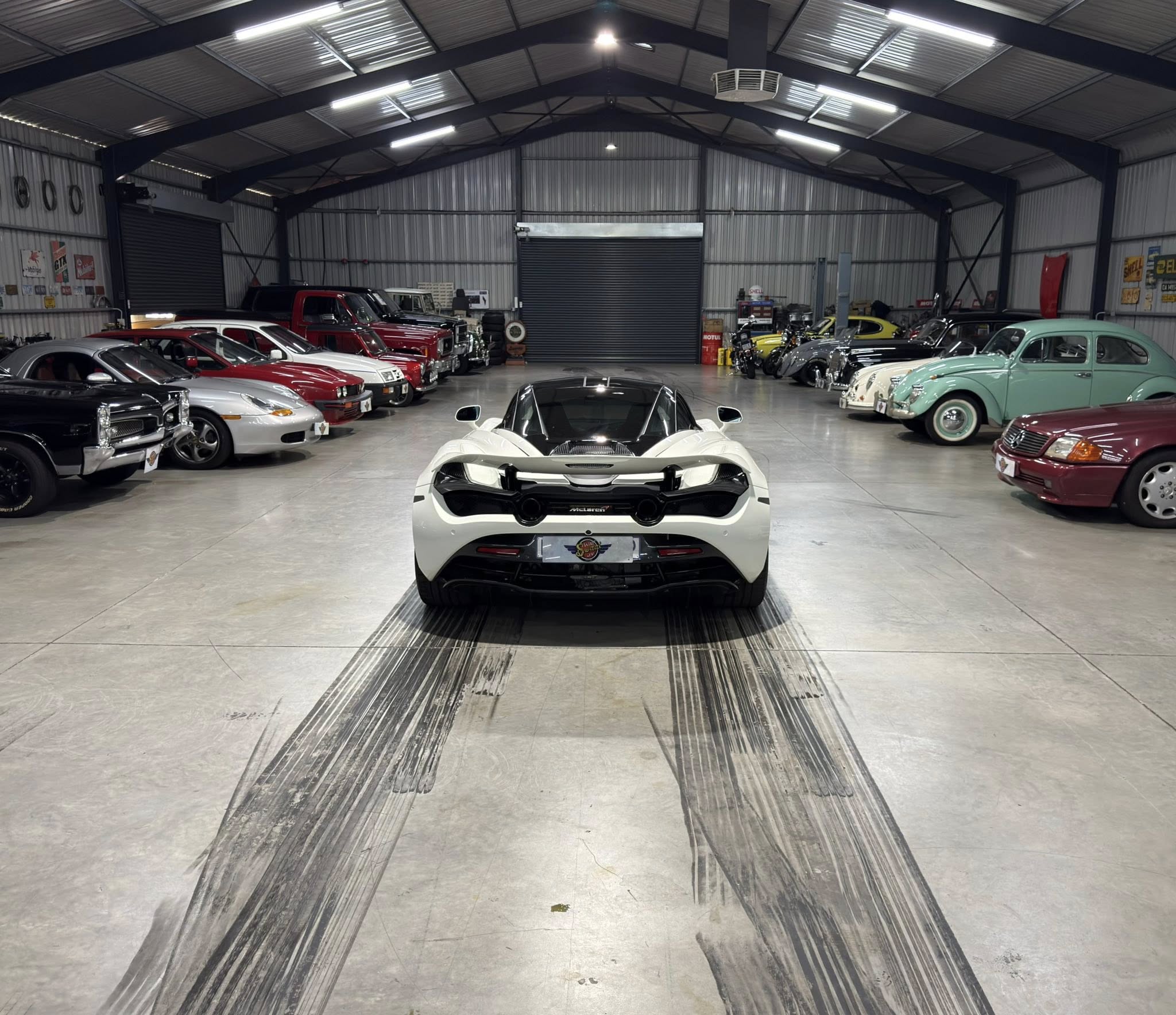 Indoor car showroom filled with classic cars along both walls, with a white McLaren centered in the foreground and a high metal ceiling above.