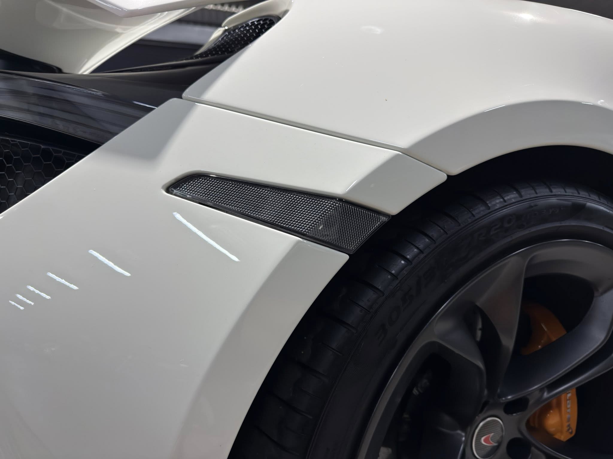 Close-up of a white sports car fender with a vent, mesh intake and visible tire edge and orange brake caliper nearby.