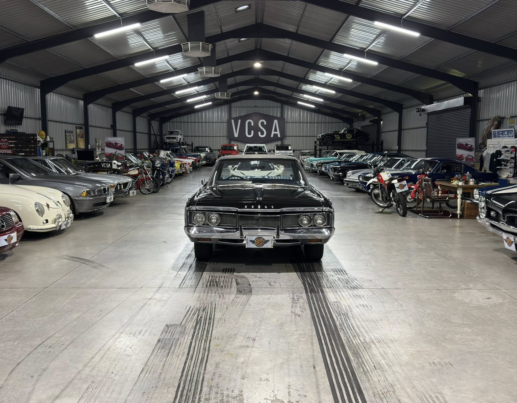 Indoor car showroom filled with classic cars lined up on both sides, facing a central black Dodge in the foreground; a large VCSA sign on the back wall under a high metal ceiling.