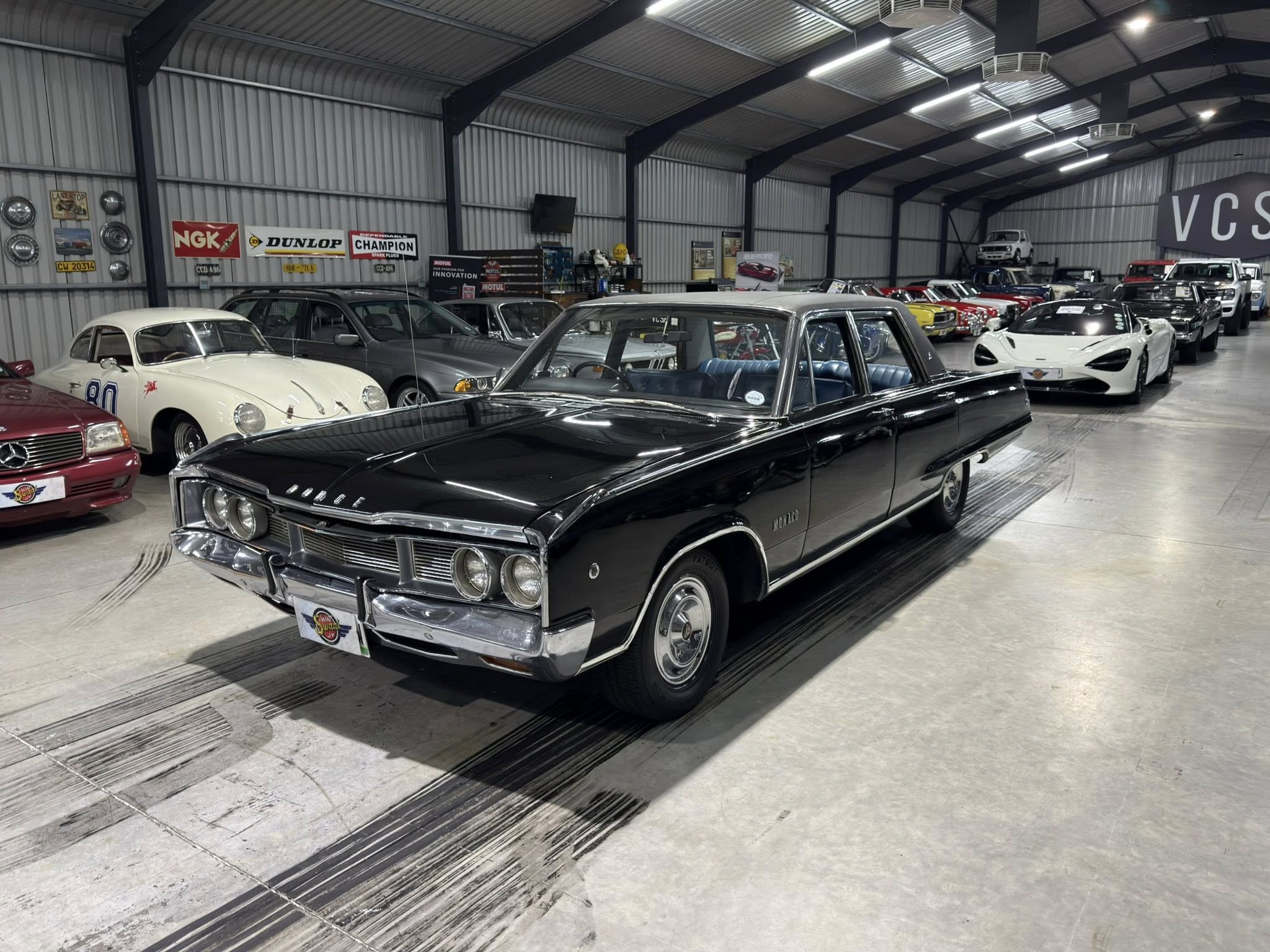 Indoor classic car showroom with vintage vehicles lined up along a metal-walled warehouse. In the foreground a black 1960s coupe.
