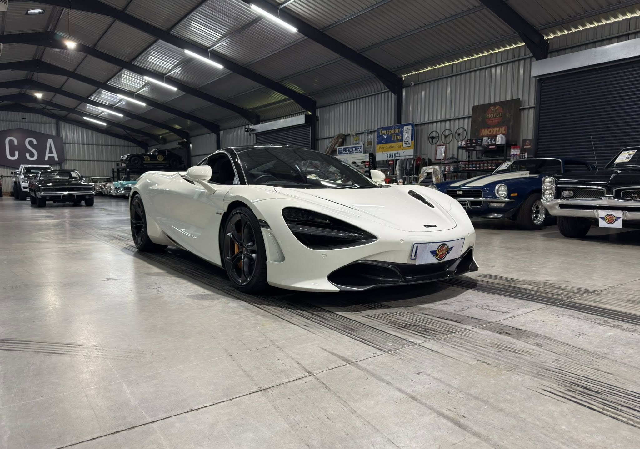 White exotic sports car on display in a large showroom surrounded by classic cars and vintage signage on the wall.