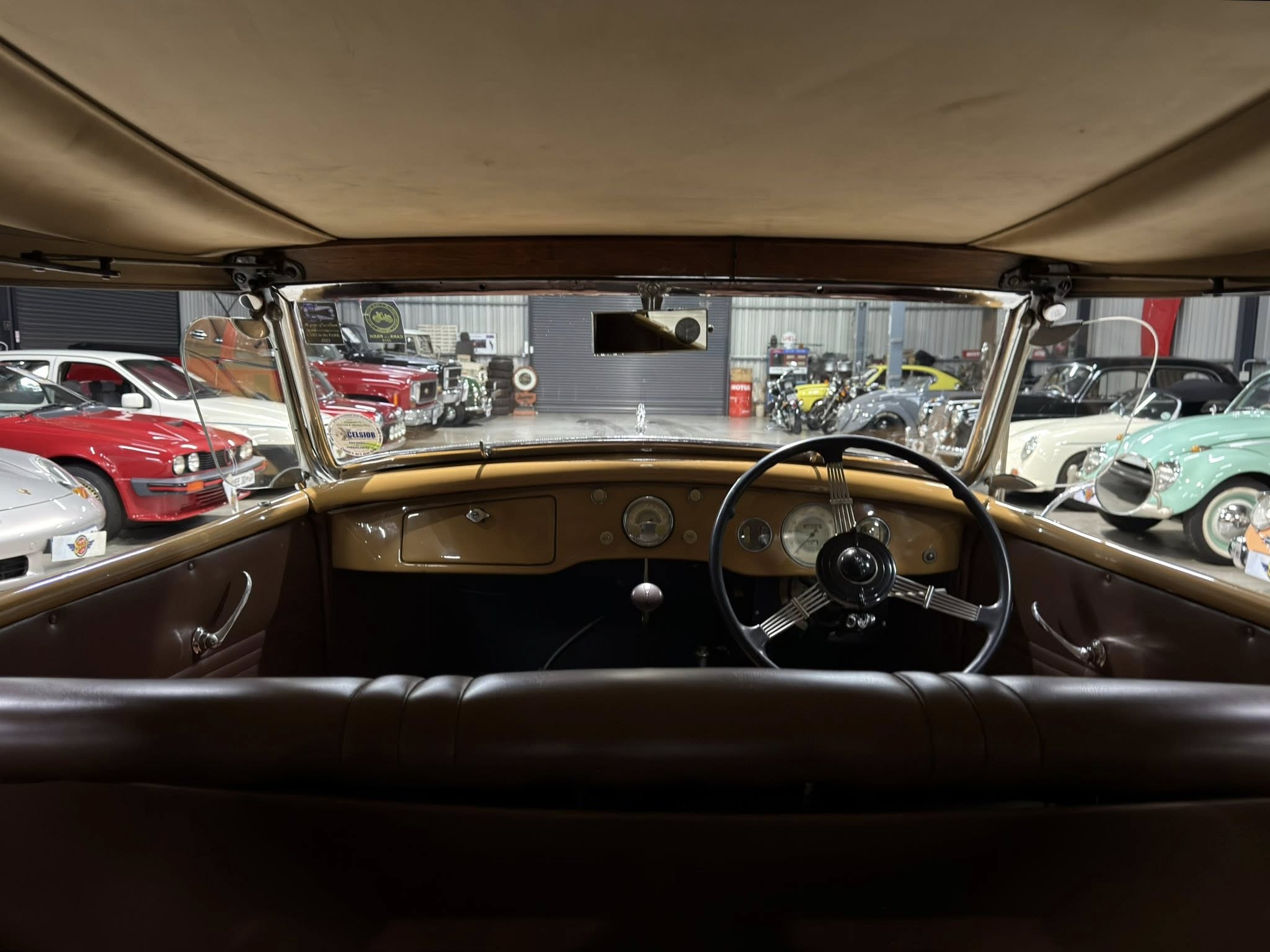 Interior view from the driver’s seat of a vintage brown car, with a showroom of classic cars visible through the windshield and side windows.