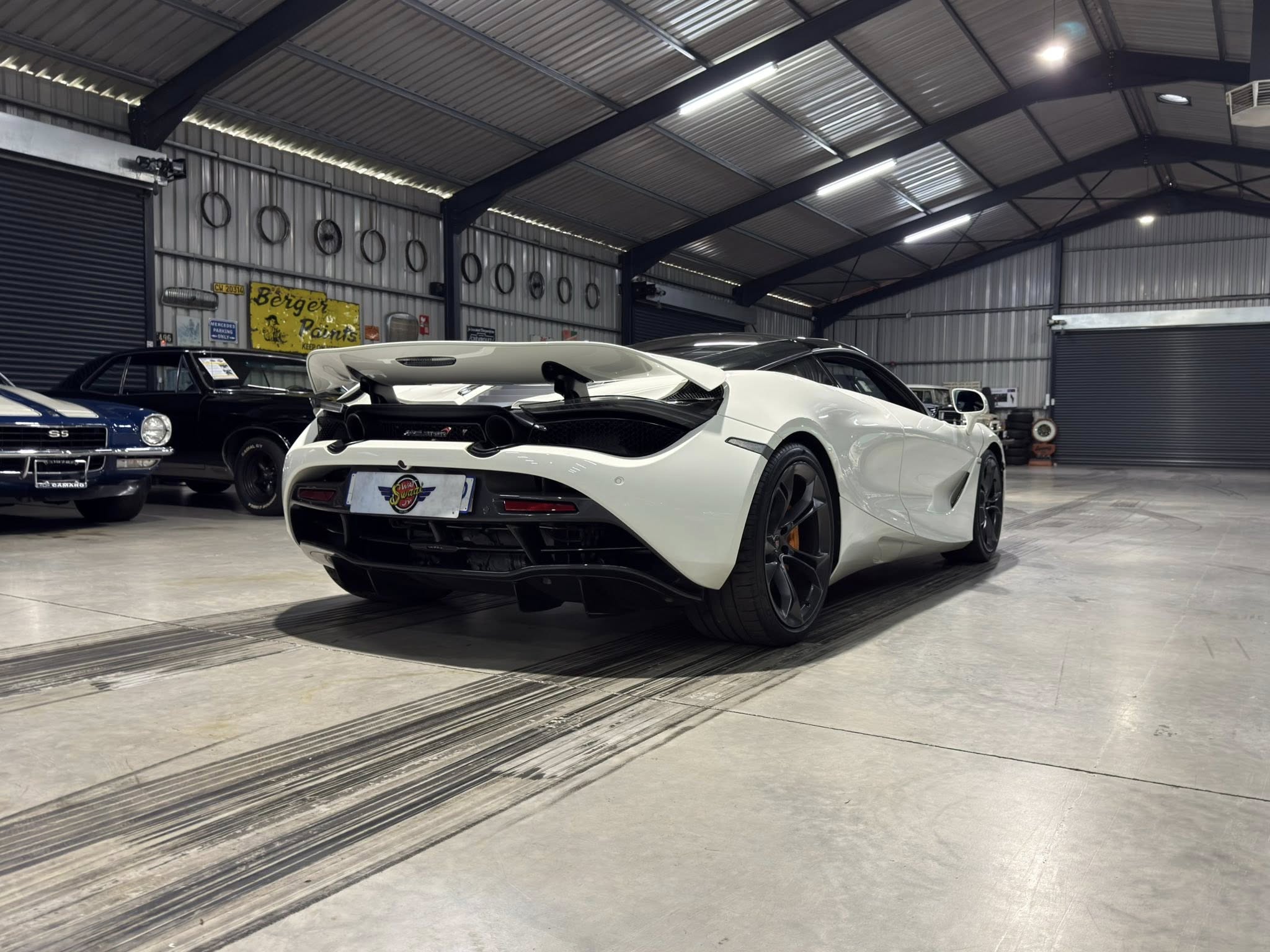 White sports car in a showroom with other classic cars along the left wall inside a metal-industrial garage.