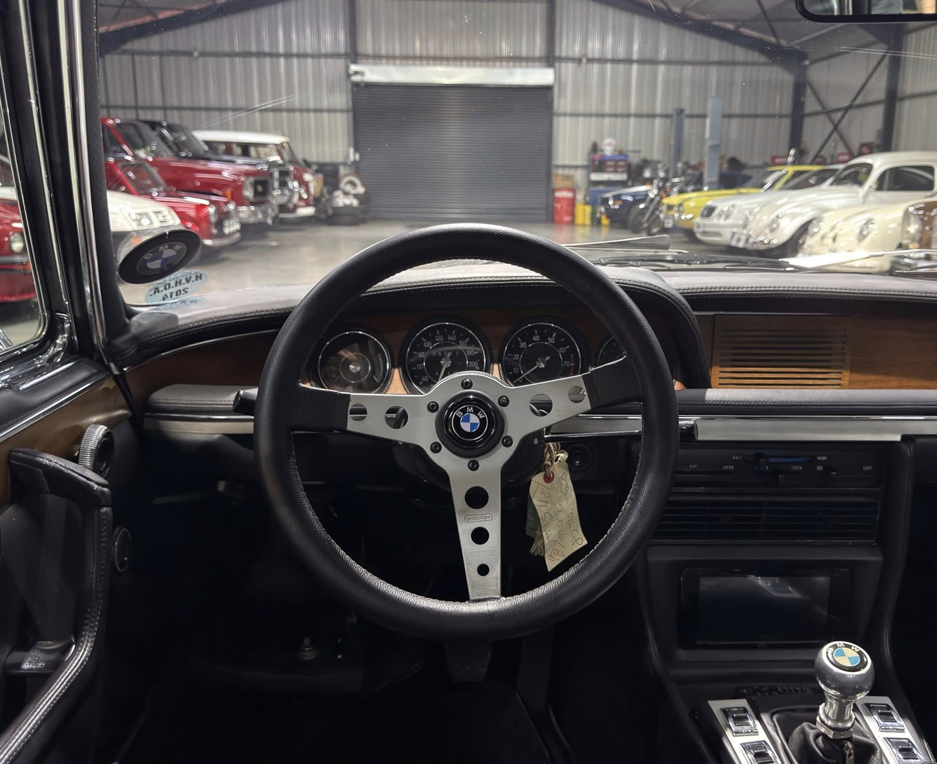 View from the driver's seat inside a vintage BMW showroom, with the classic steering wheel and dashboard, and rows of old cars outside.