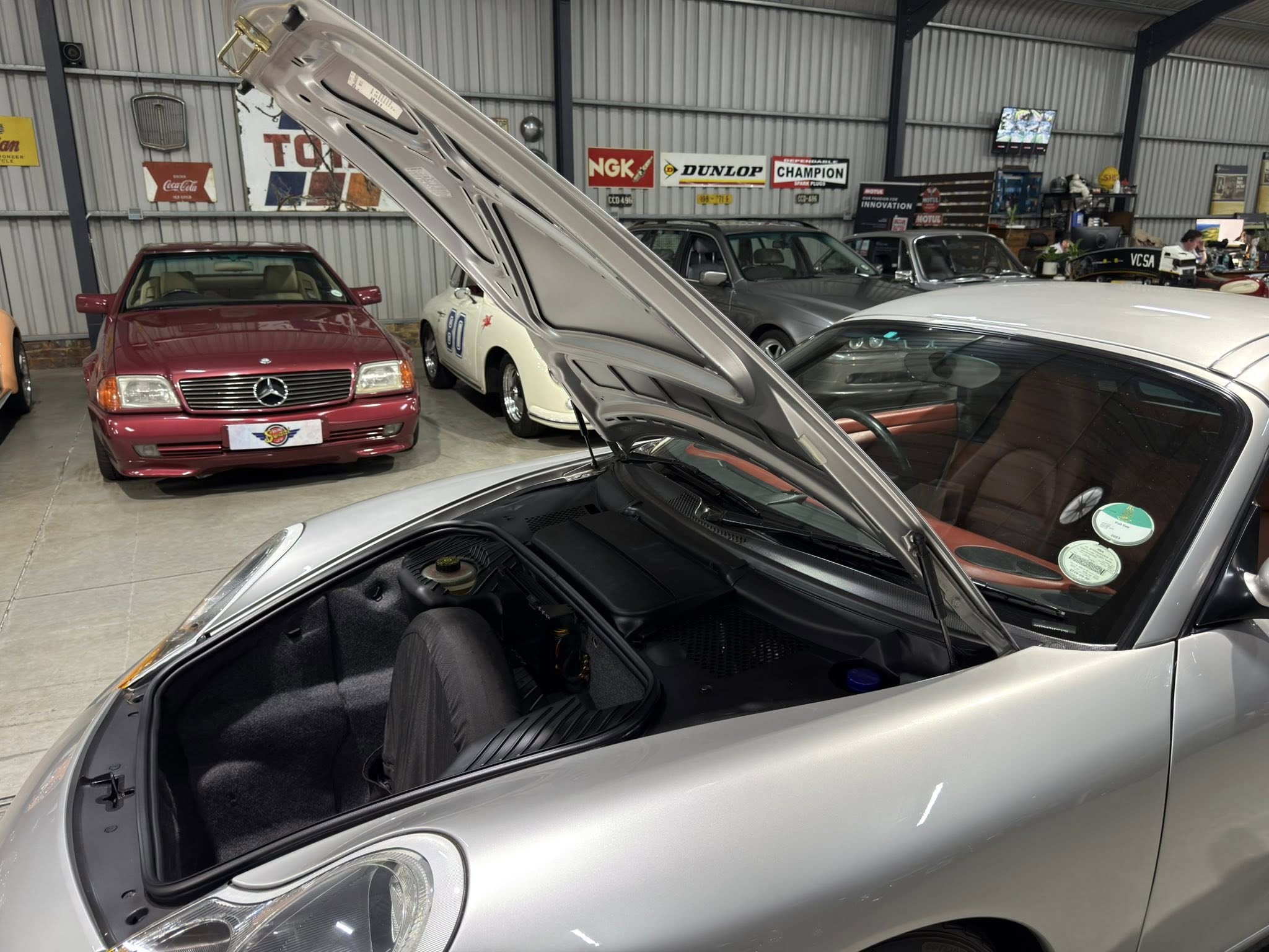 Silver car with its hood raised in a garage showroom, other classic cars visible in the background under metal walls with banners.