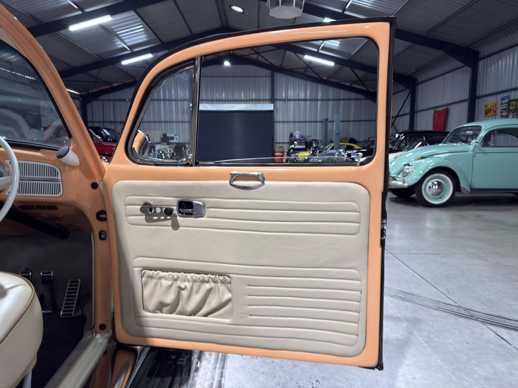 Open peach-colored car door revealing tan interior upholstery in a showroom full of classic cars; teal and other vintage cars visible in the background.