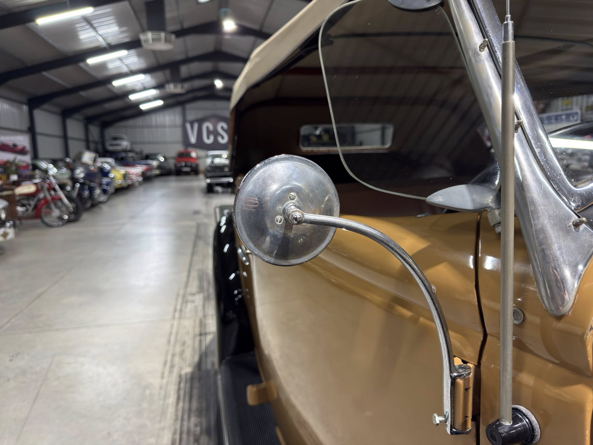 Close-up of chrome side mirror and bumper on a vintage car inside a workshop, with other vehicles blurred in the background and a 'VCS' sign visible.
