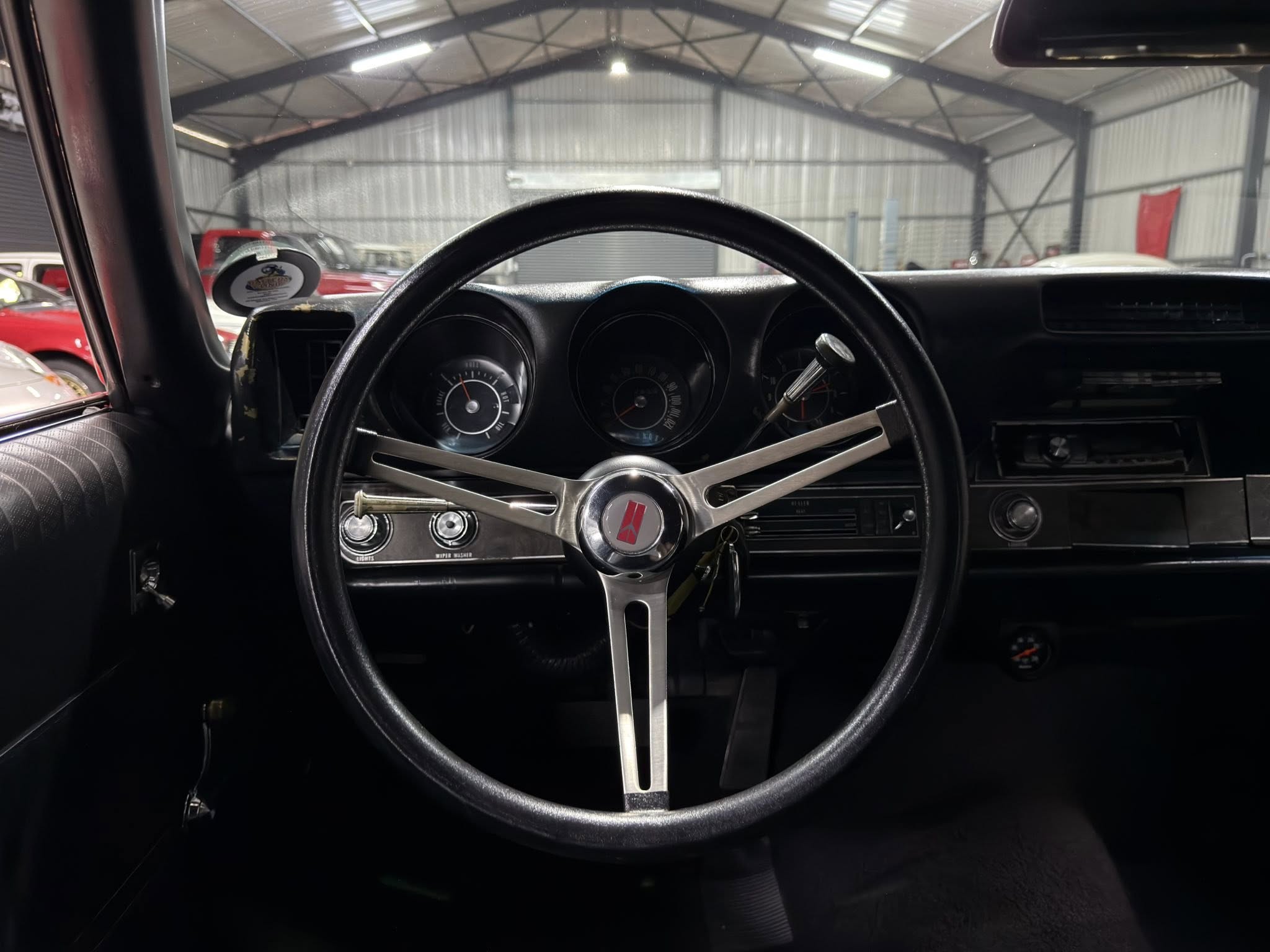 View from the driver's seat of a vintage car, showing a three-spoke chrome steering wheel and classic dashboard in a dim garage.