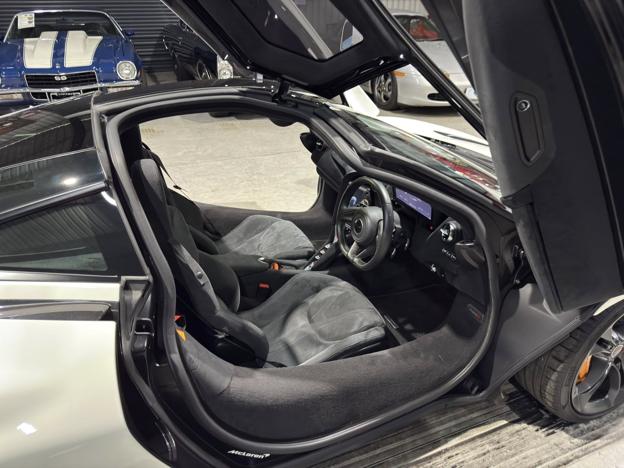Open McLaren interior with door raised, showing black seats, steering wheel, and center console; blue carorso background in a showroom.