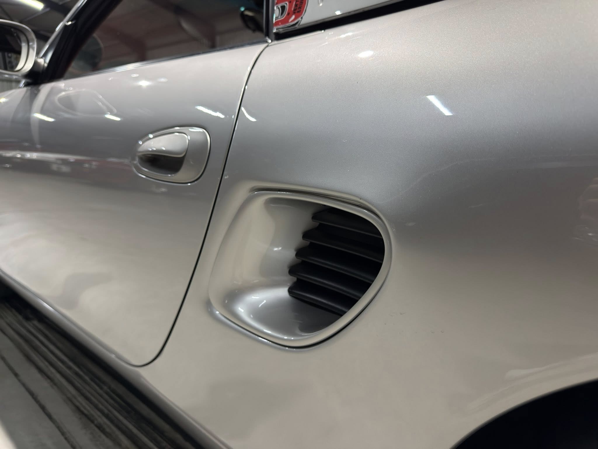 Close-up of a silver car’s side panel showing the door handle, vent, and glossy metallic finish in a showroom light reflection.