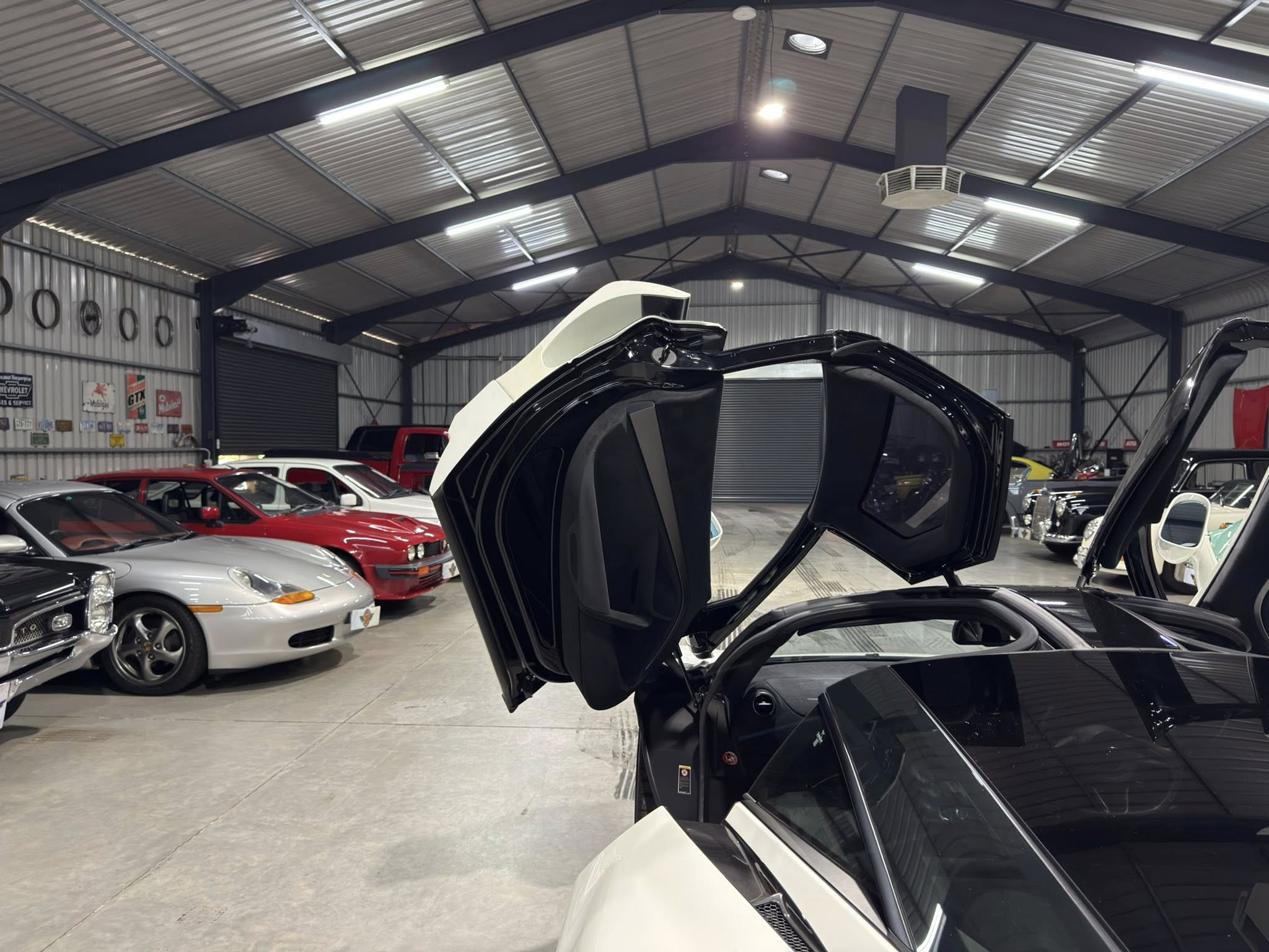 Inside a large showroom or garage with several cars, a black luxury car has its gull-wing doors open in the foreground.