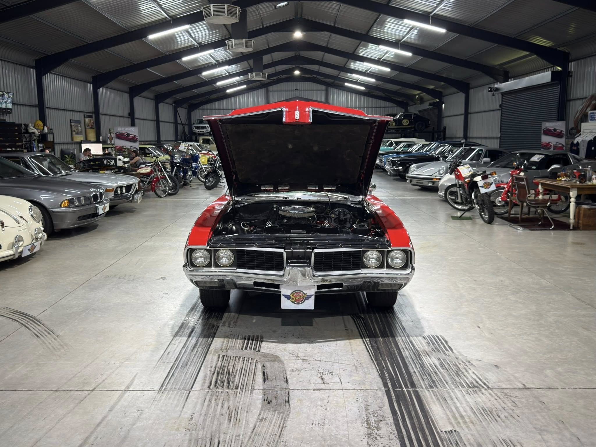 Red classic car with its hood open in a large showroom, surrounded by other cars and motorcycles on display.