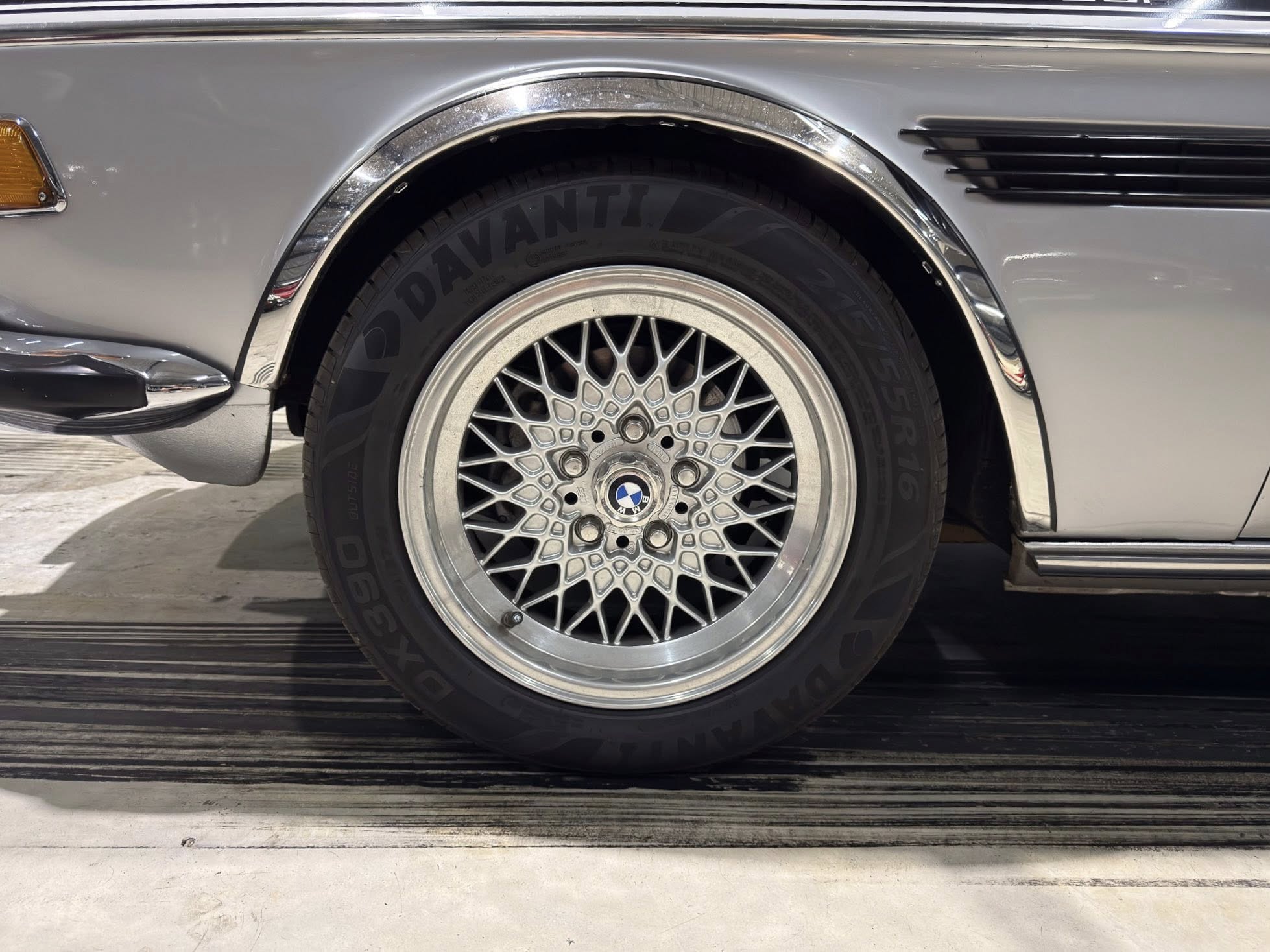 Close-up of a silver car's front wheel with a detailed mesh alloy rim and chrome fender arch nearby.