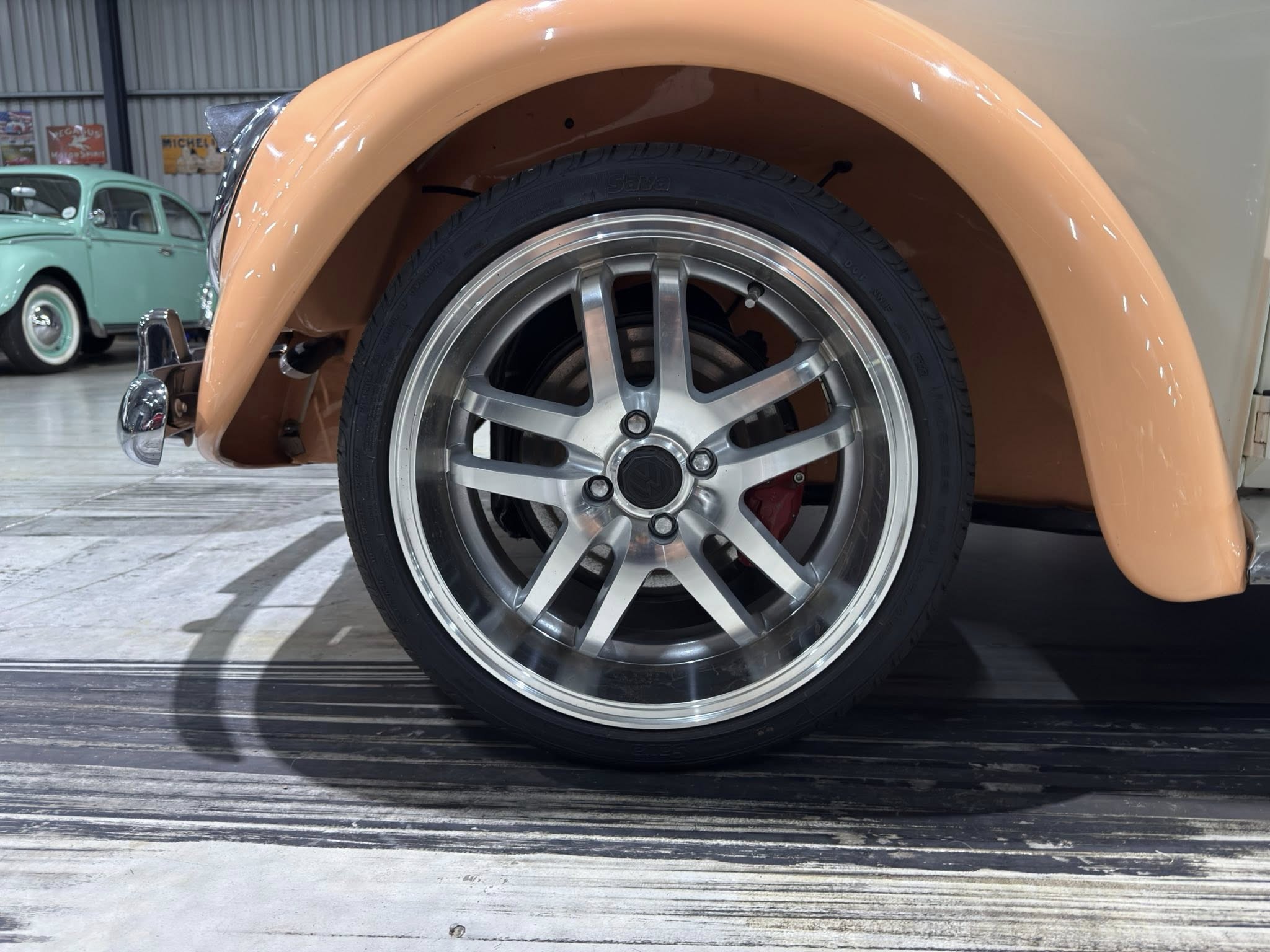 Close-up of a car wheel with a silver rim under a peach fender in a showroom with a vintage green car in the background.