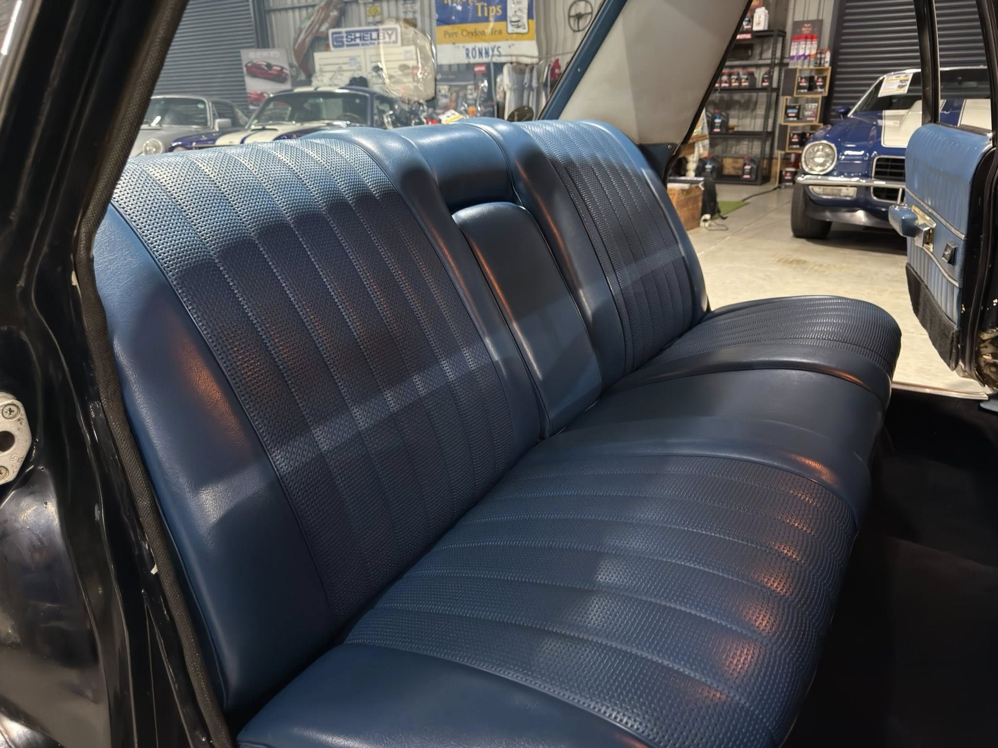 Blue rear bench seat in a classic car, open door revealing a showroom with vintage cars in the background.