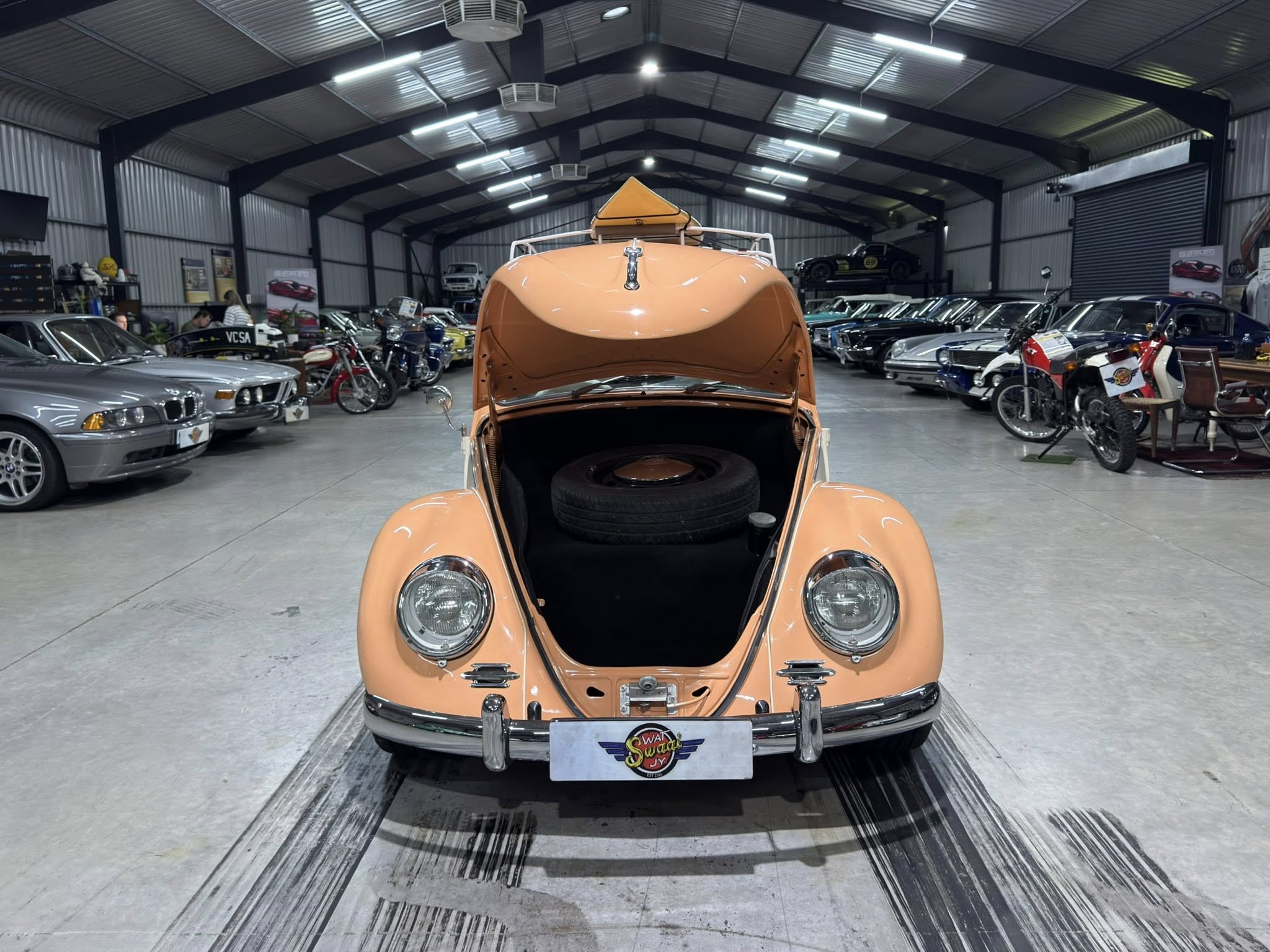 Orange vintage car with open hood showcased in a large showroom filled with classic cars and motorcycles in the background.