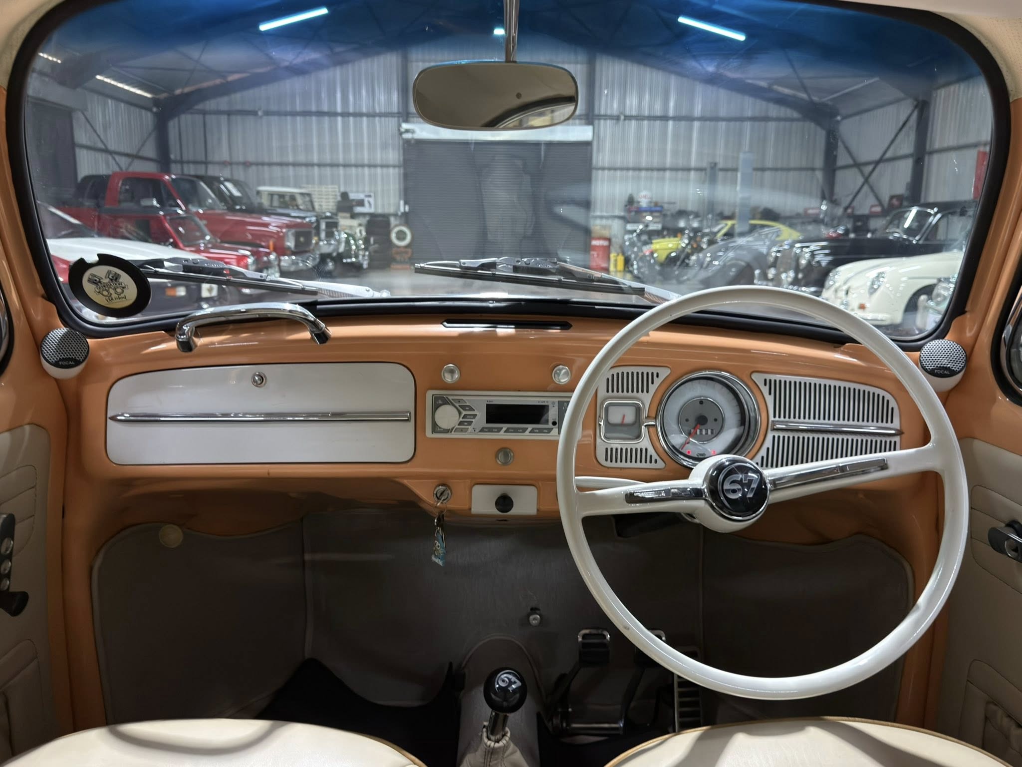 From the driver's seat: vintage orange car interior with white steering wheel and dashboard, looking out at a garage full of classic cars.