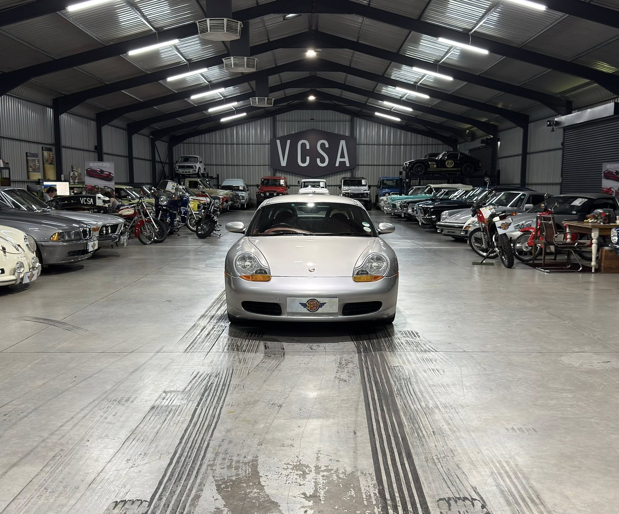 Indoor showroom filled with classic cars, with a prominent VCSA sign at the back center. A silver sports car sits in the foreground.