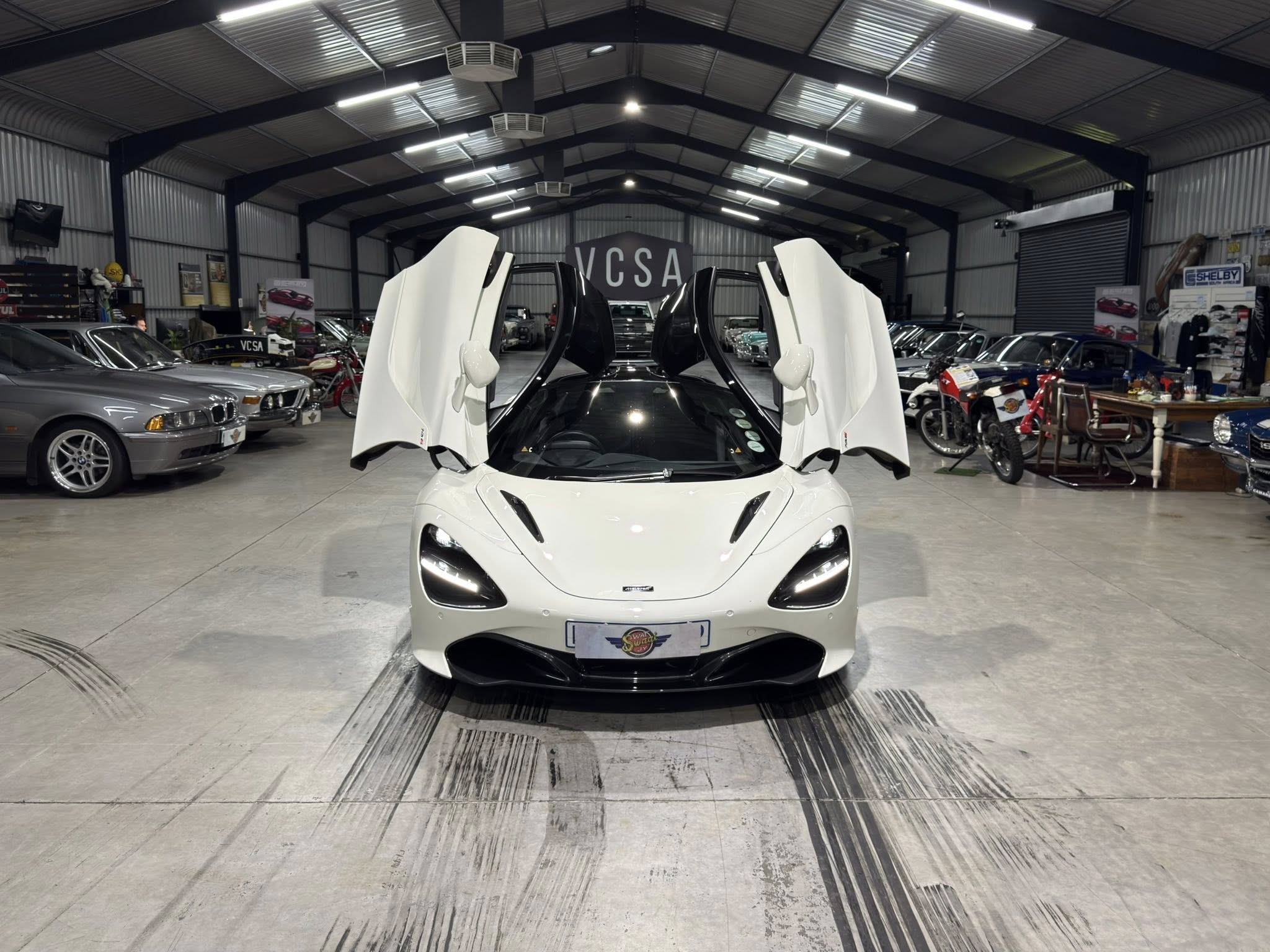 White sports car with open butterfly doors inside a showroom, other vehicles and posters in the background.
