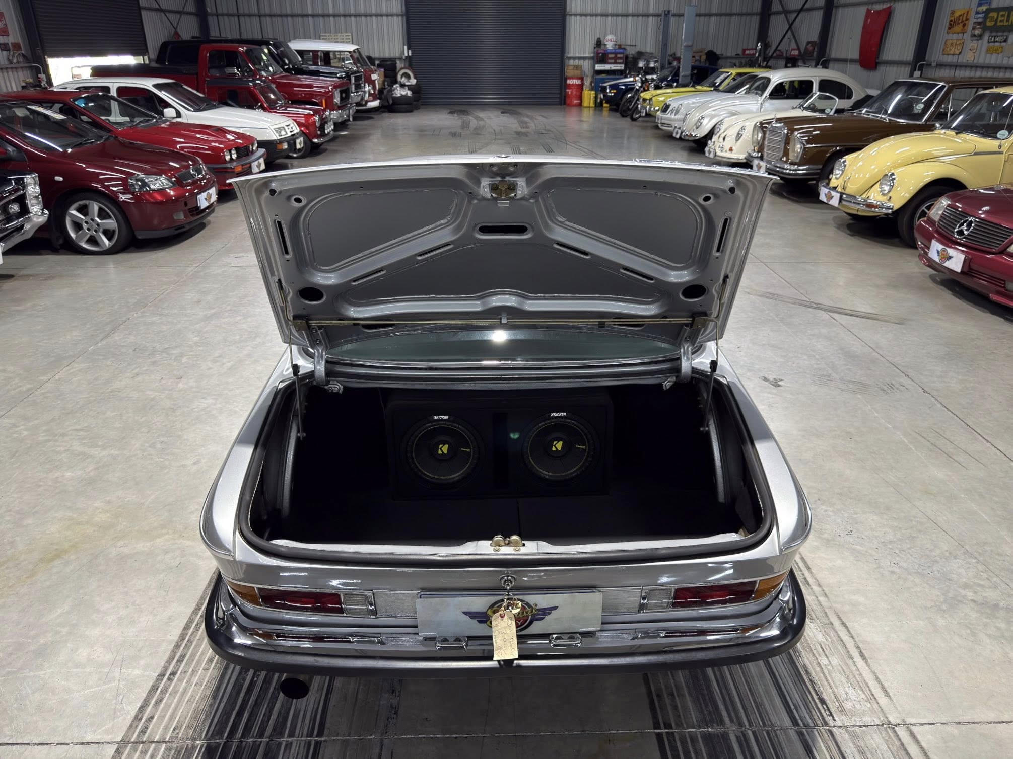 Open trunk of a silver car in a showroom lined with vintage cars on both sides.