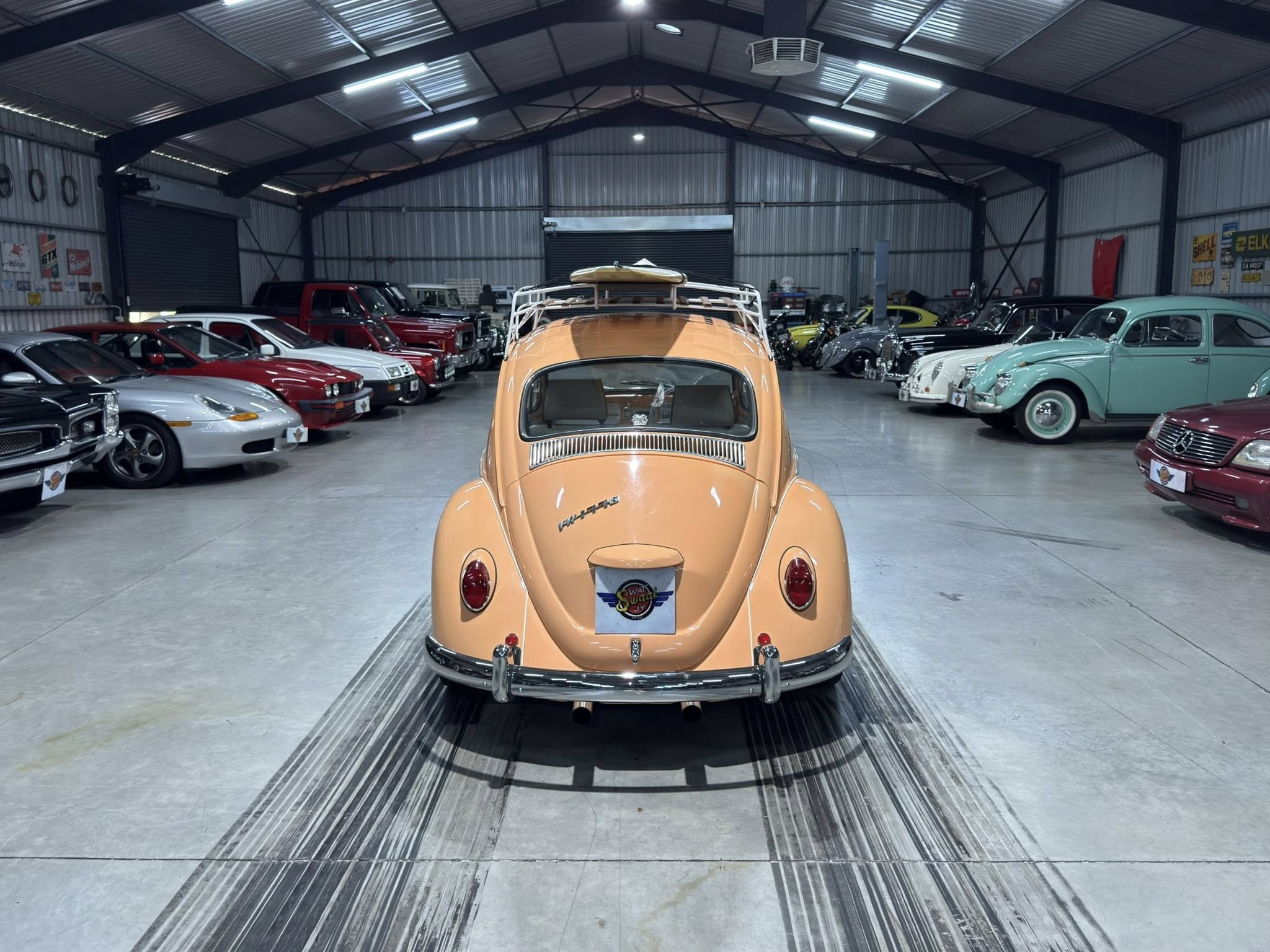 Orange vintage Volkswagen Beetle viewed from the rear in a showroom filled with classic cars on both sides, with a surfboard on its roof rack.