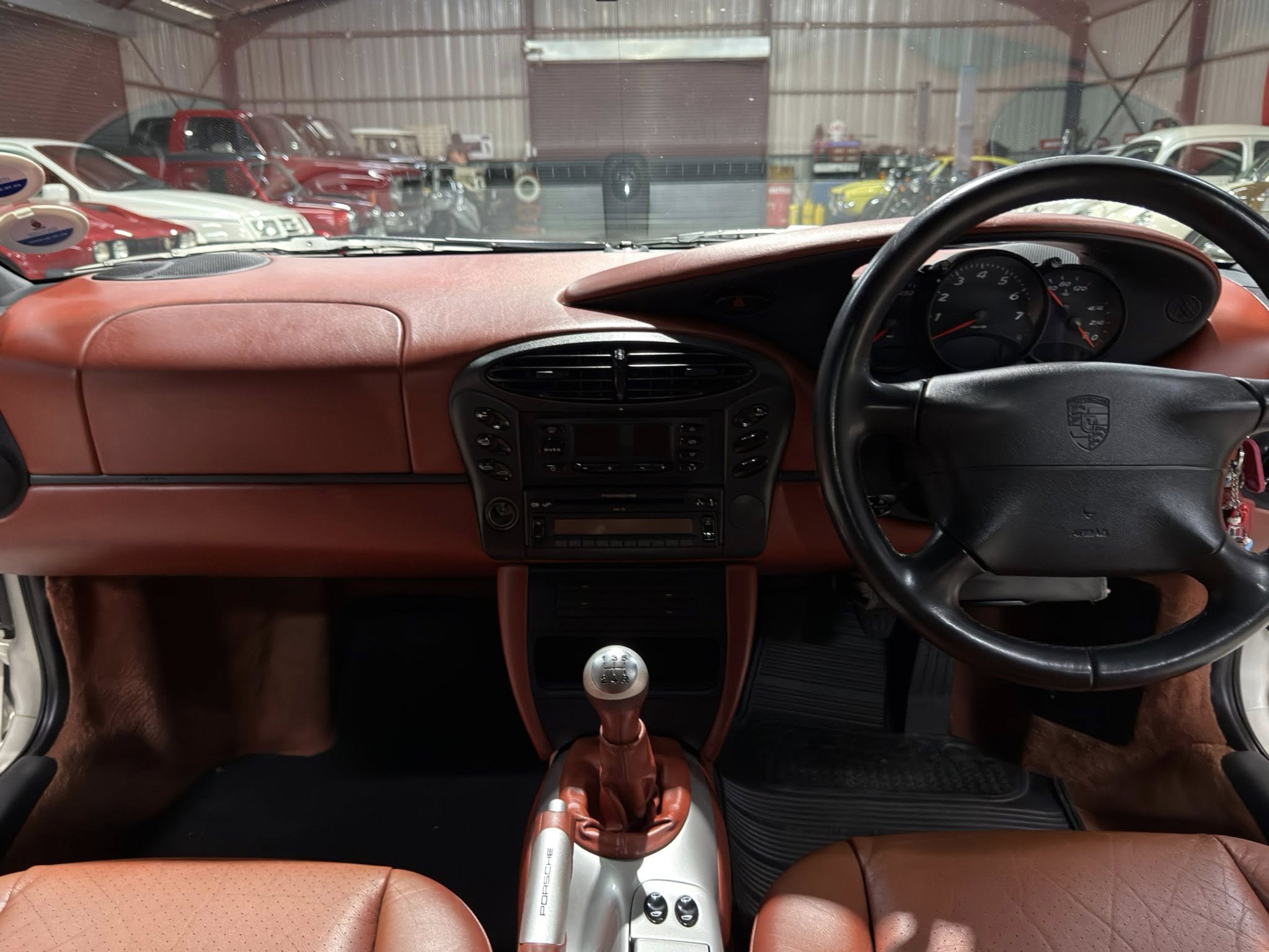 Inside a red-car cockpit with a manual shifter, steering wheel, and tan leather seats; view through the windshield of a workshop with parked cars in the background