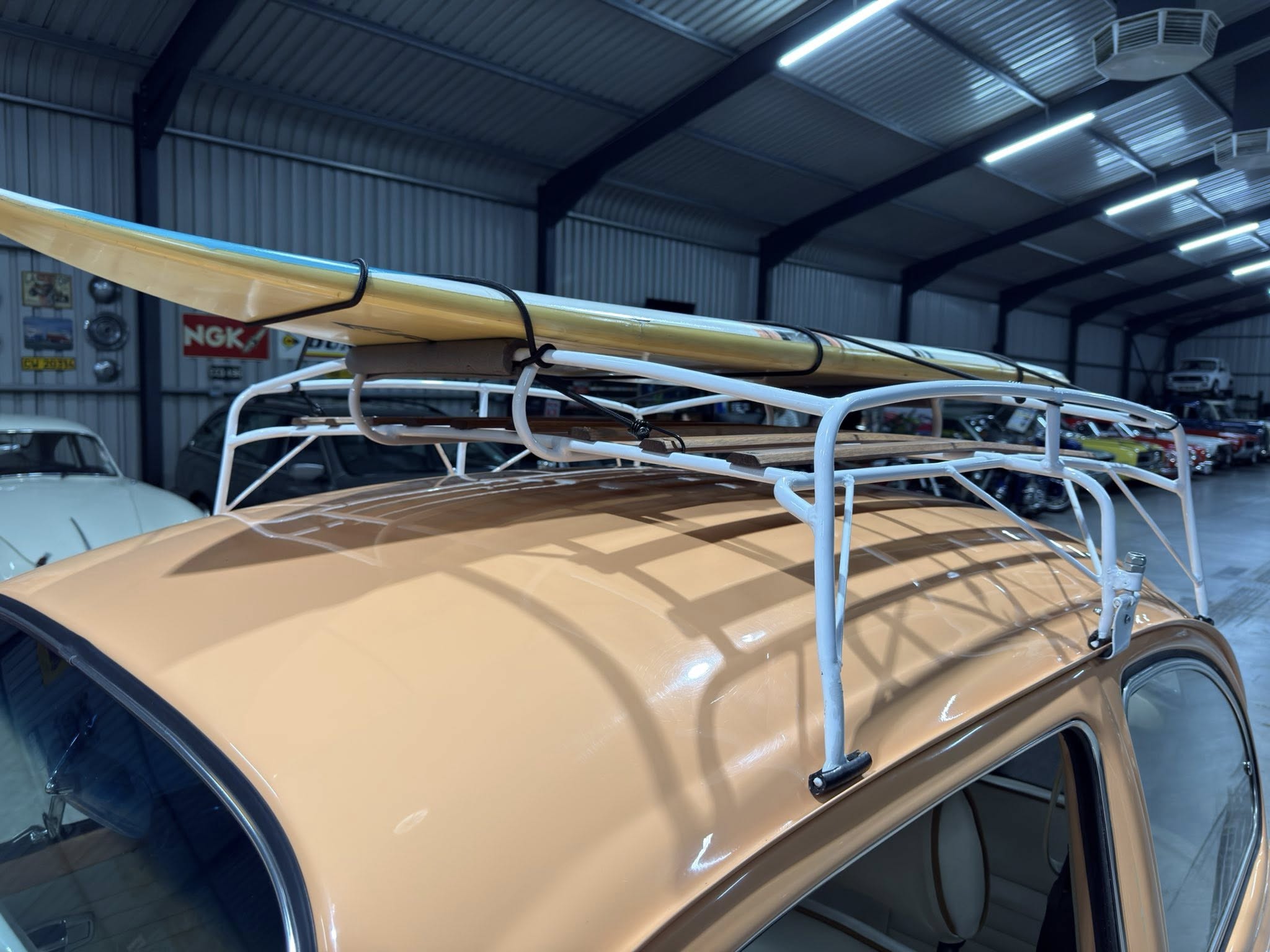 Beige car indoors with a yellow surfboard strapped to a roof rack in a workshop/garage.