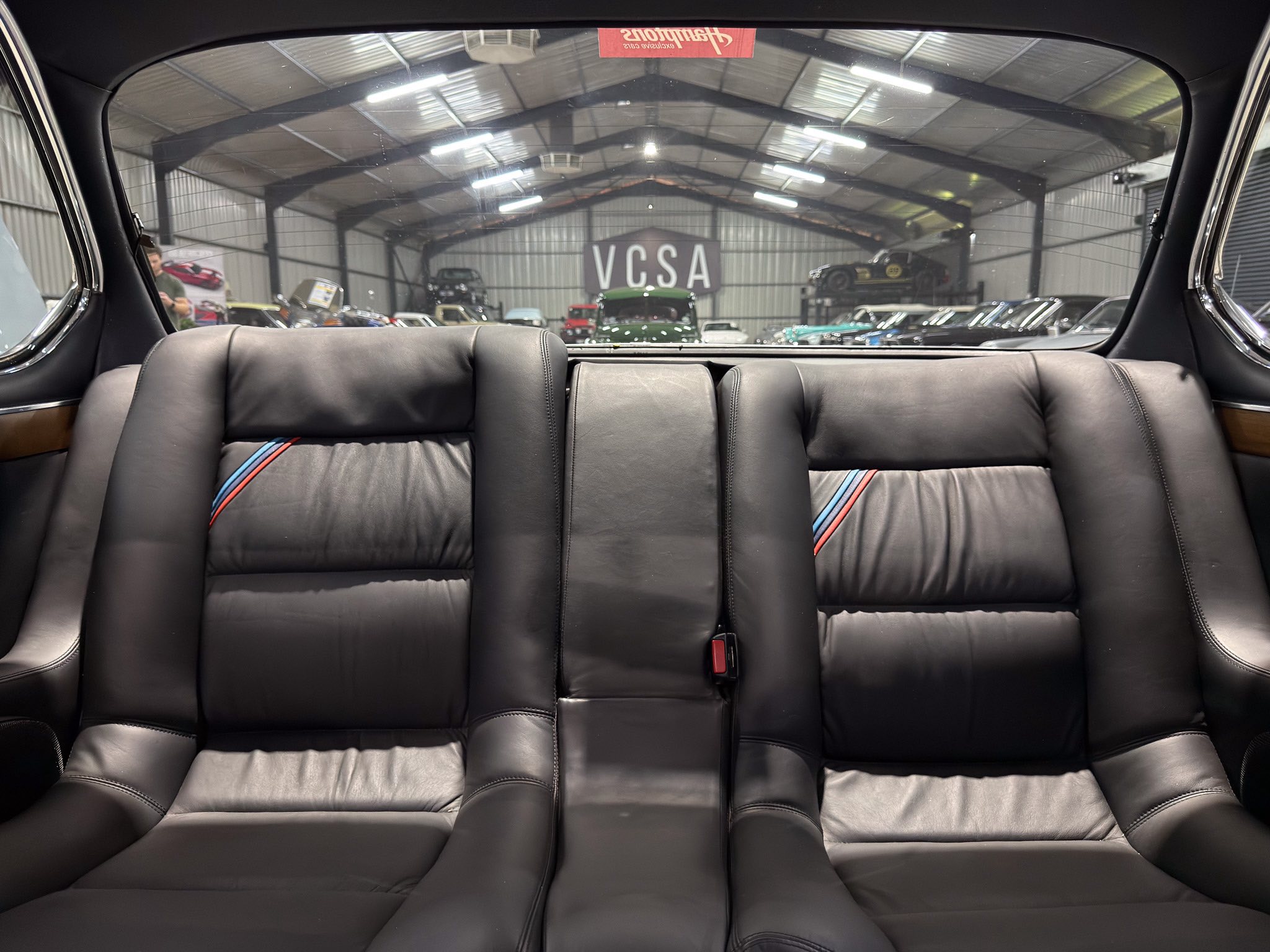 View from the back seat of a car looking out through the rear window at an indoor car showroom with multiple vehicles on display and a VCSA sign in the distance.