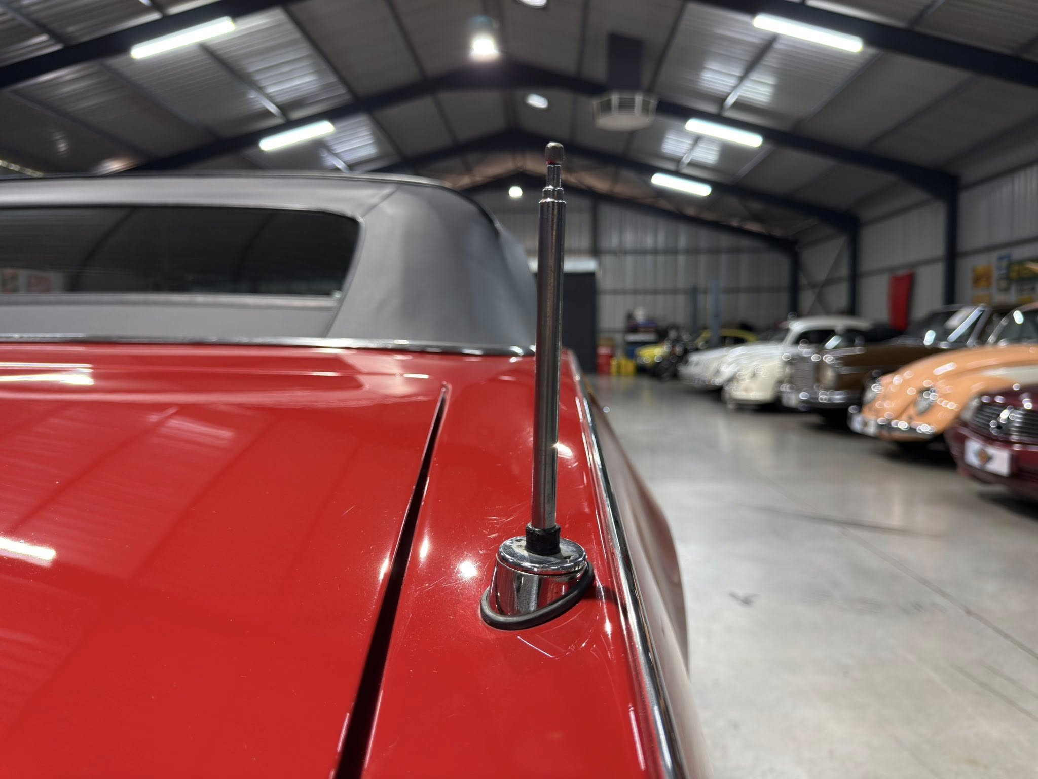Close-up of a car antenna mounted on a red roof in a large indoor showroom or garage, with vintage cars in the background.