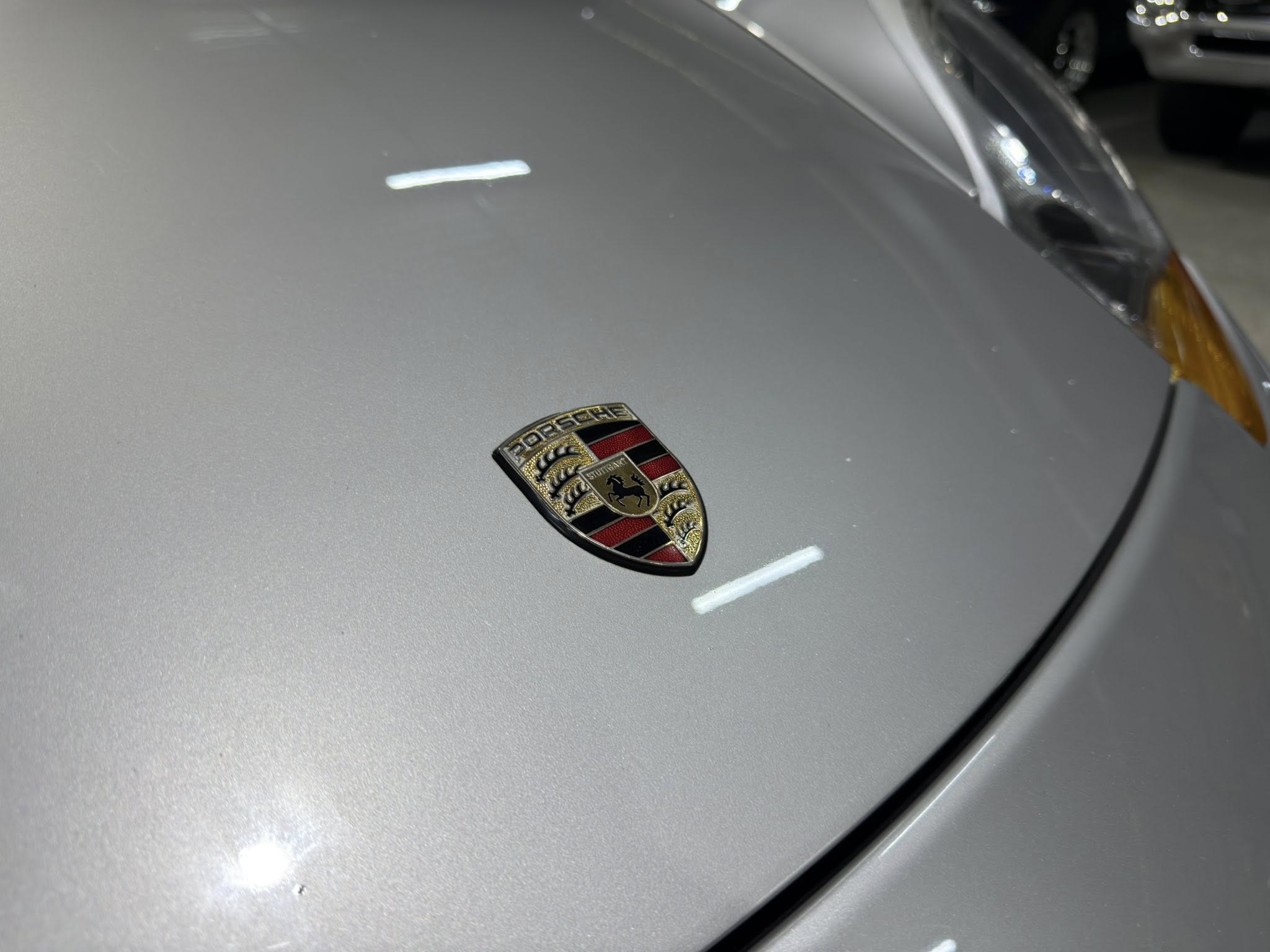 Close-up of a silver Porsche hood with the emblem in the foreground and the right-side headlight partially visible in the background.