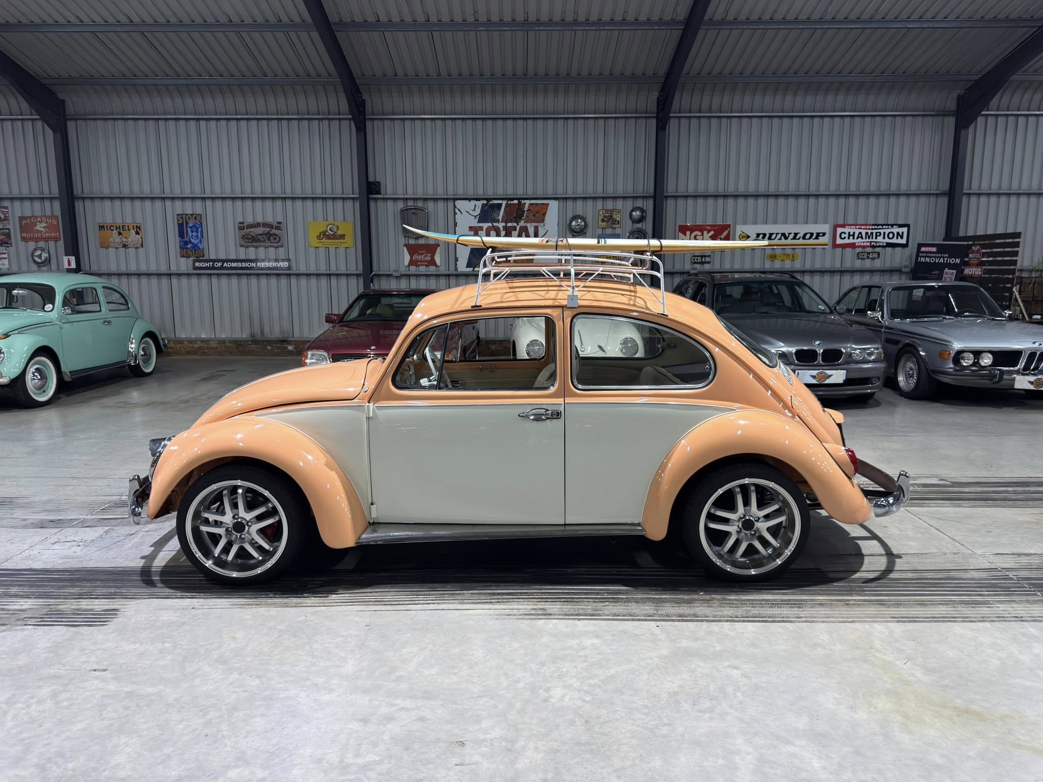 Orange vintage car with a roof rack in a showroom, other classic cars and retro signage on the back wall behind it.