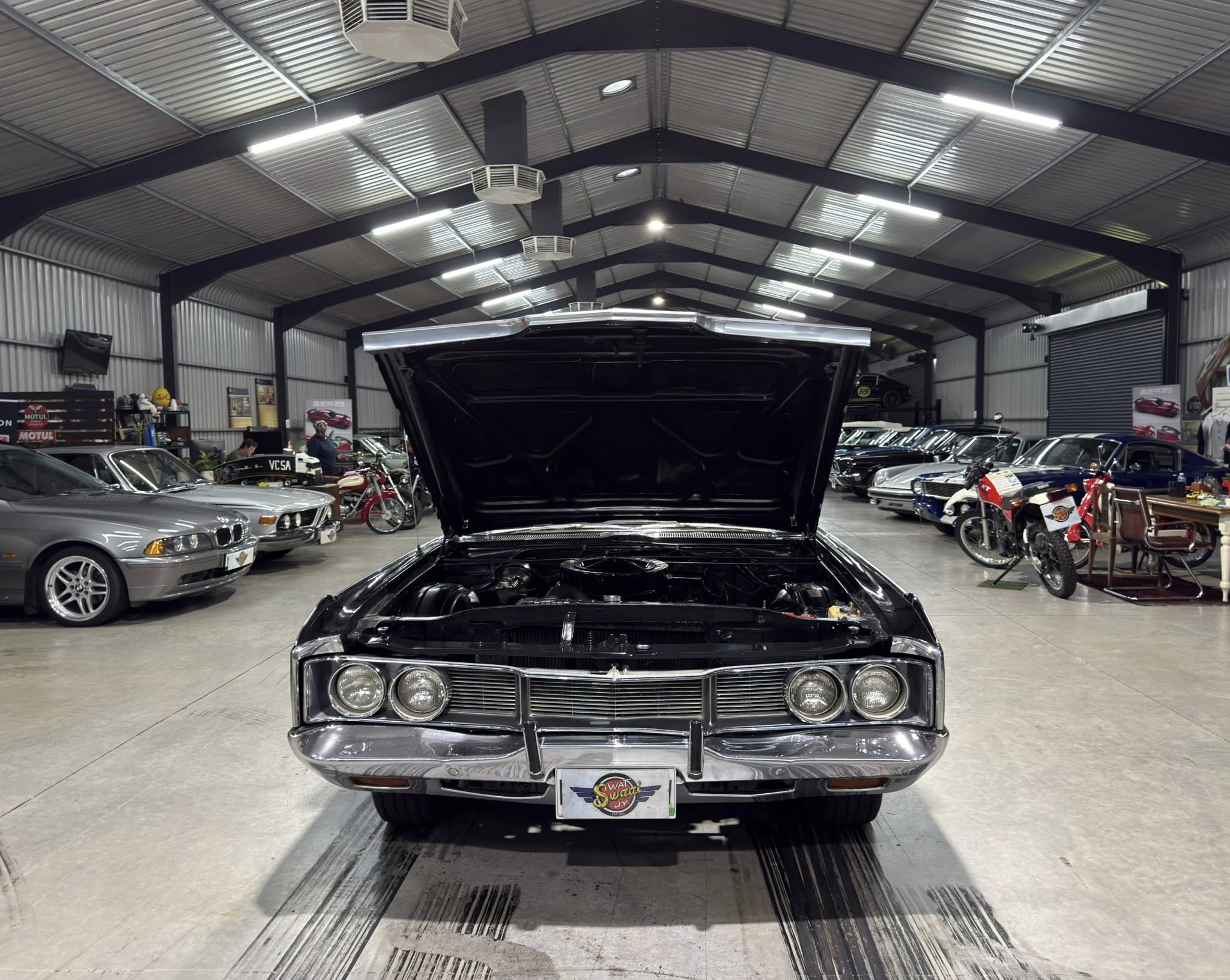Front view of a black vintage car with its hood open inside a well-lit car showroom filled with classic cars and motorcycles in the background, ceiling with metal beams above.