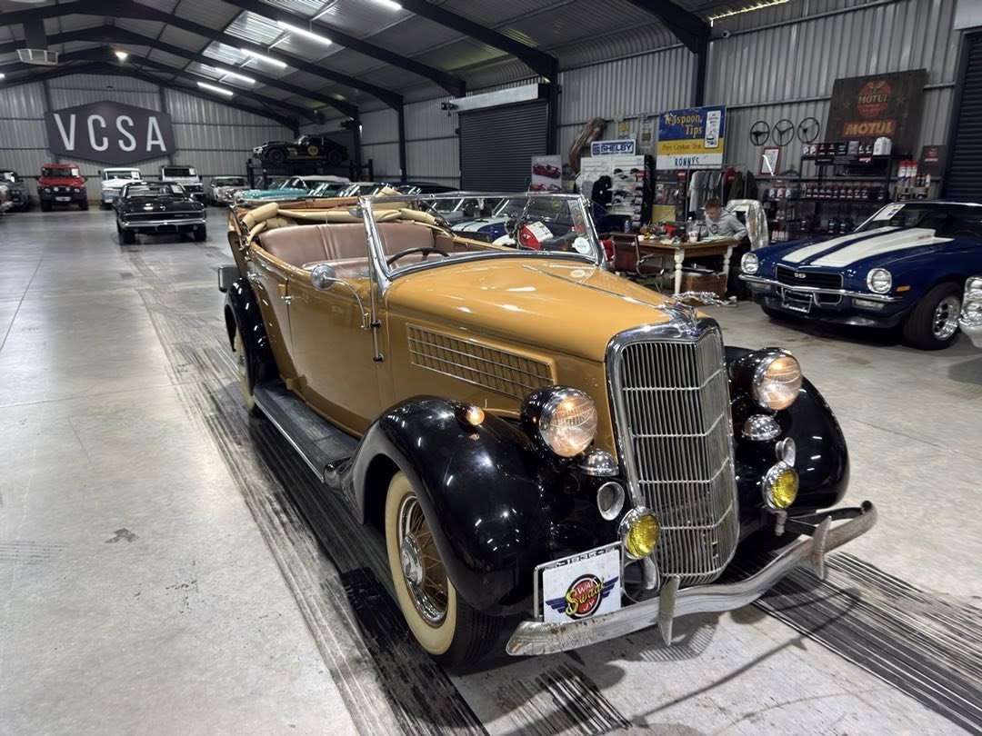 A tan and black vintage convertible car inside a showroom, with other classic cars in the background and a large 'VCSA' sign on the wall.