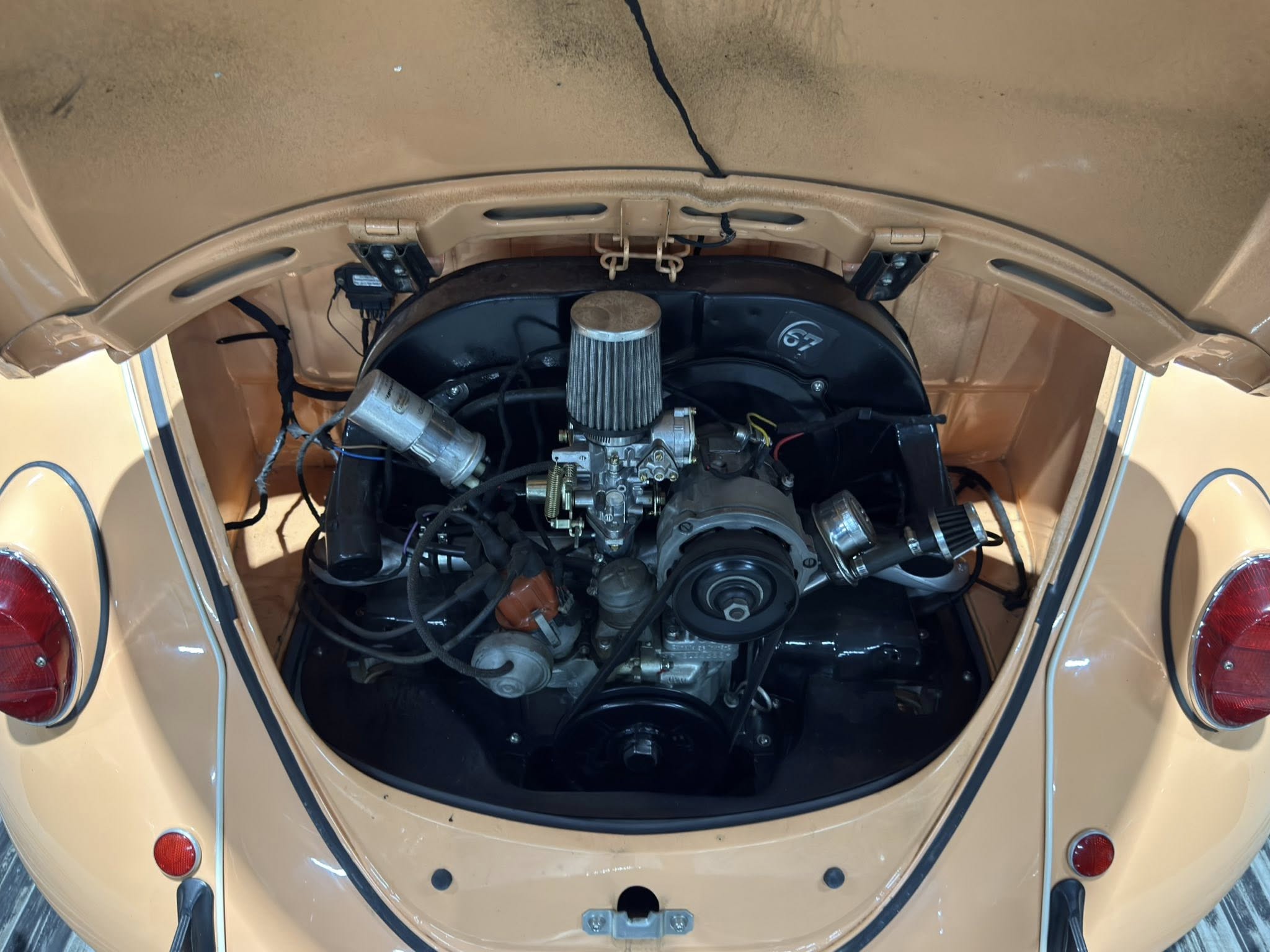Rear engine bay of a vintage beige car, showing the carburetor and large air filter with visible wiring and hoses.