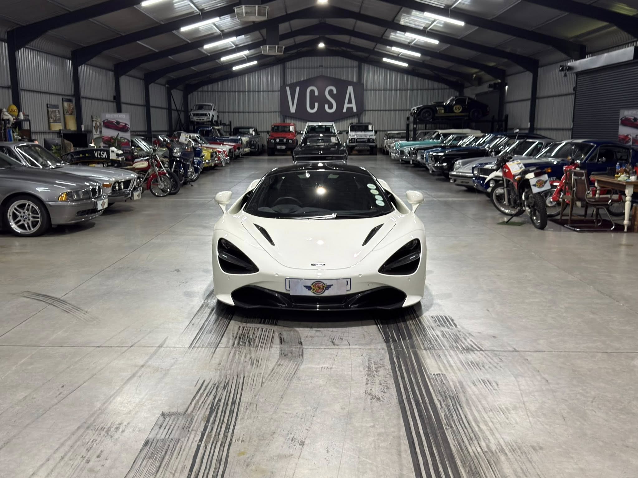 Interior of a car showroom with rows of vintage and modern cars, a white sports car centered in the foreground and a large 'VCSA' sign at the back.