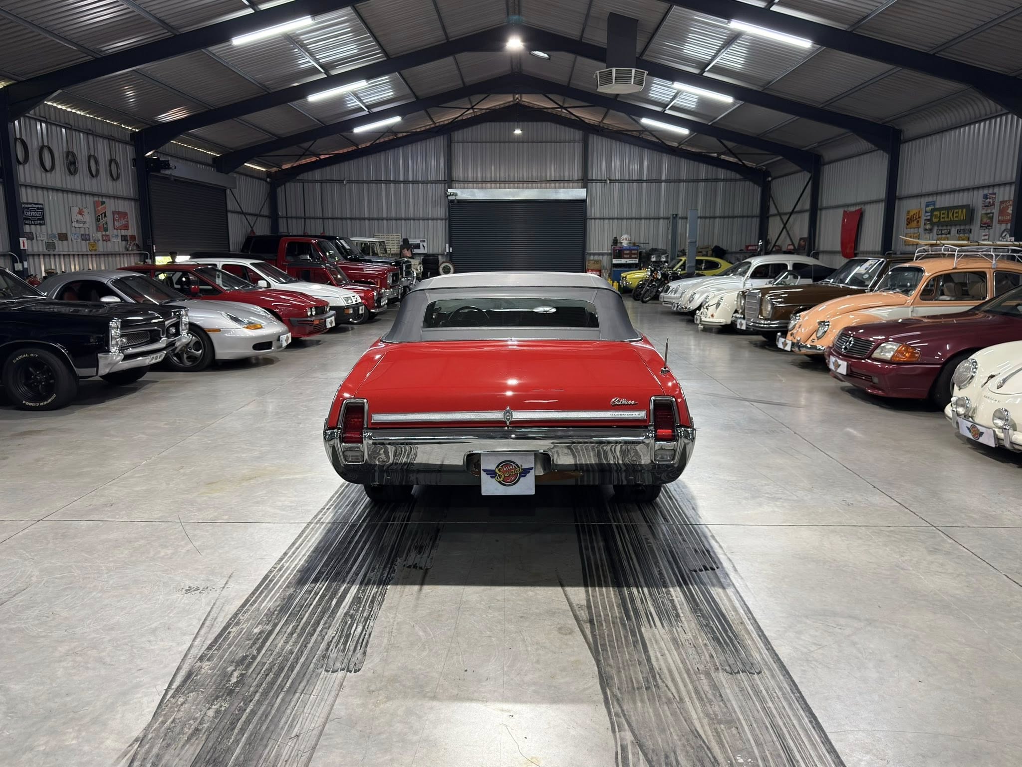 Indoor showroom filled with vintage cars lined along both sides of a wide central aisle in a metal‑walled garage.