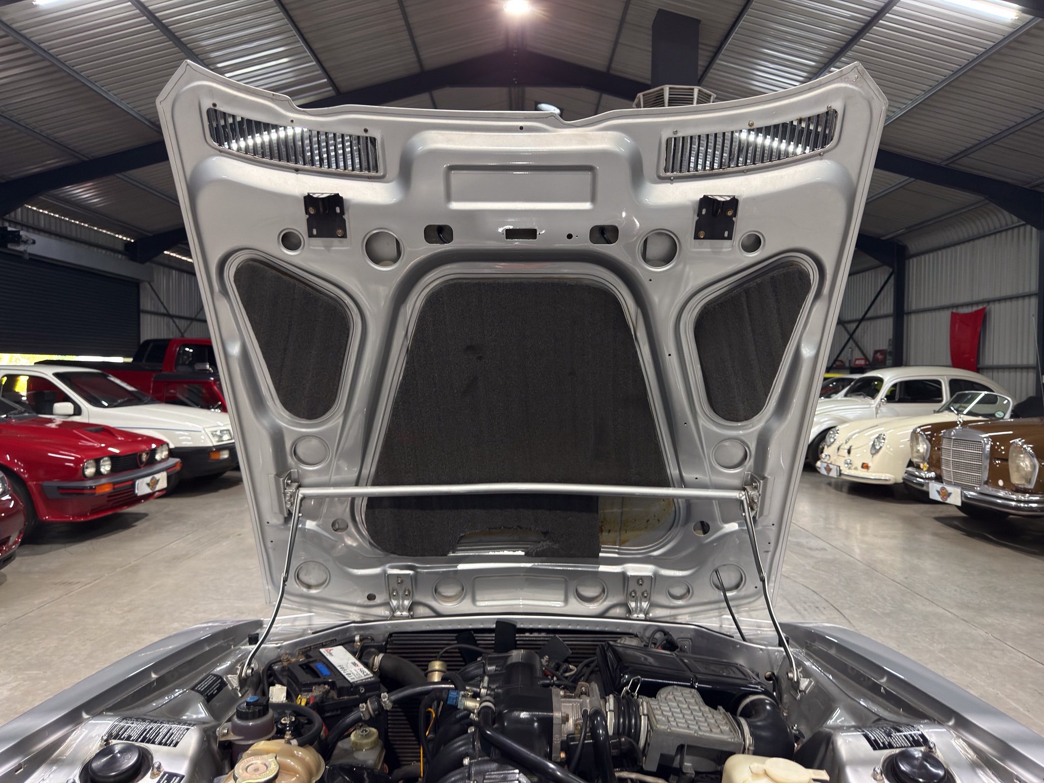Hood of a silver car raised in a showroom, exposing the engine bay with other classic cars visible in the background