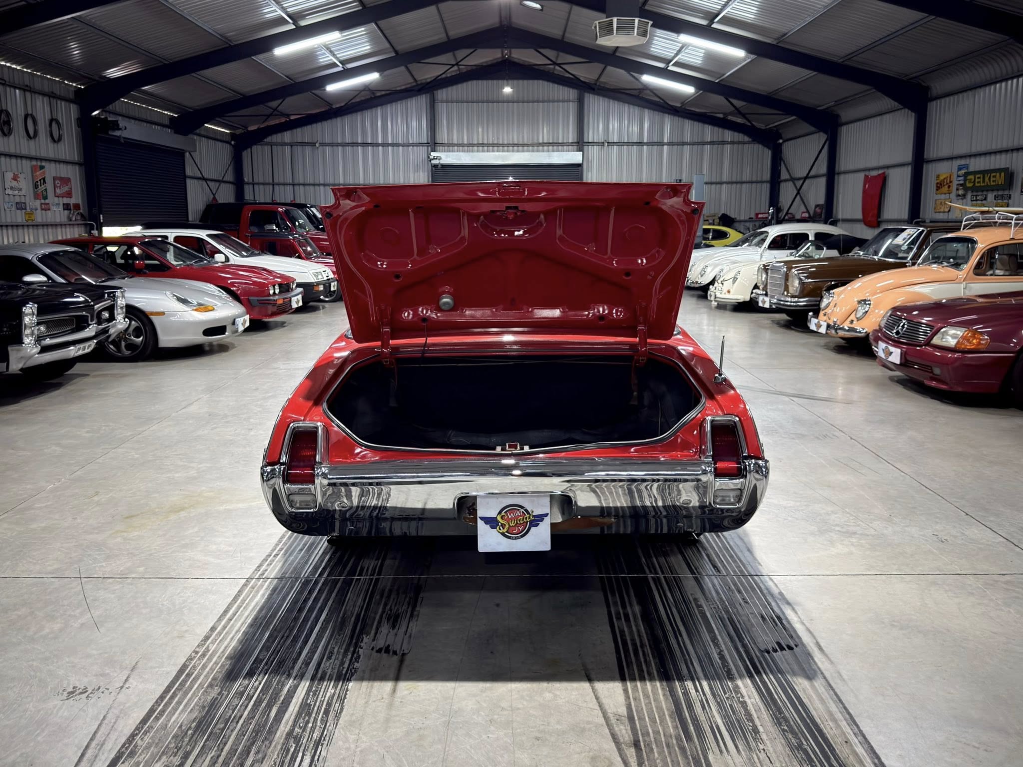 Red classic car with its trunk open in a showroom filled with vintage cars inside a large warehouse-like space, bright lighting above.